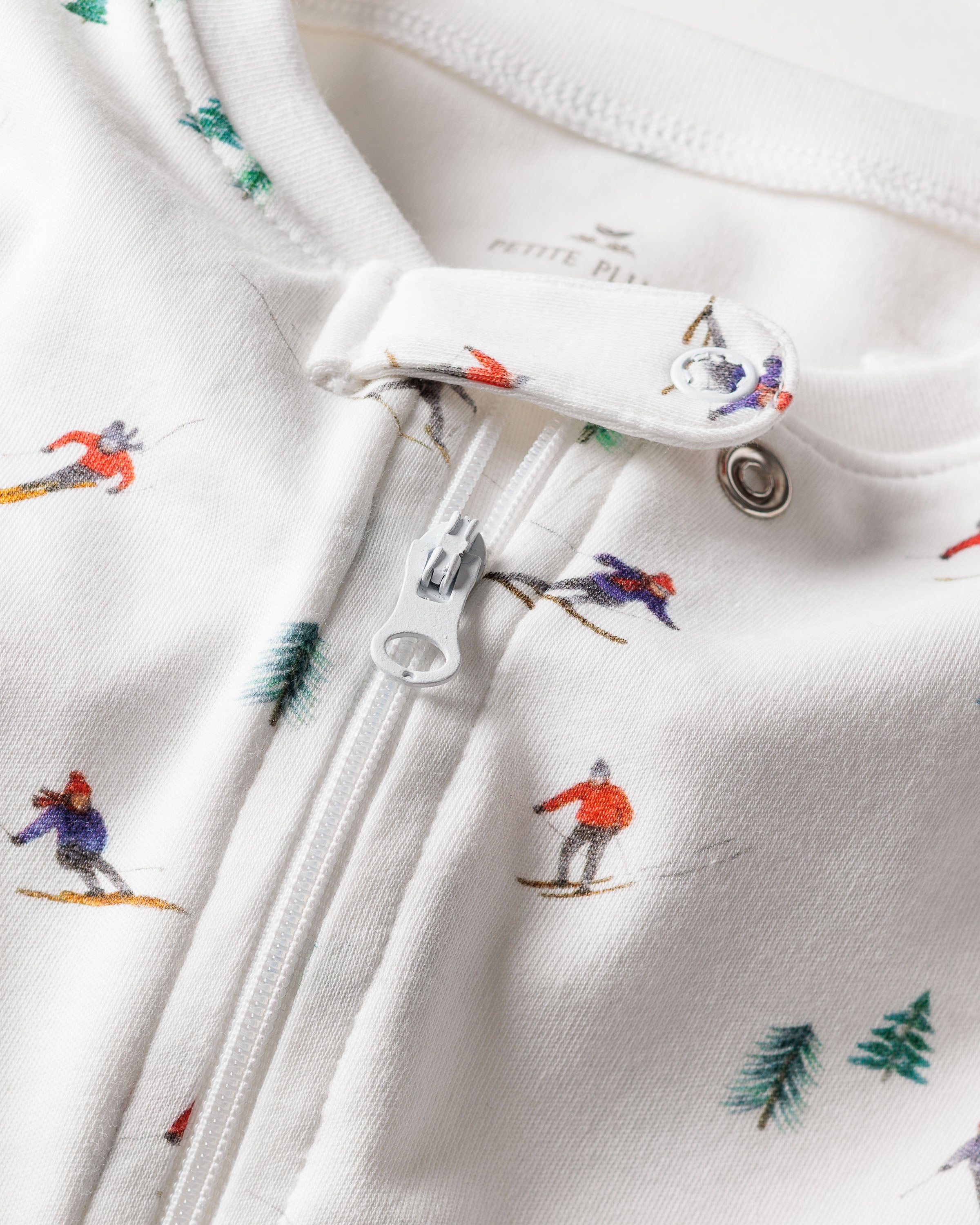 Close-up of Petite Plumes Babys Pima Romper in Après Ski, showcasing a cozy fabric with a zipper adorned with small skier illustrations and scattered green trees, ideal for sleepwear.
