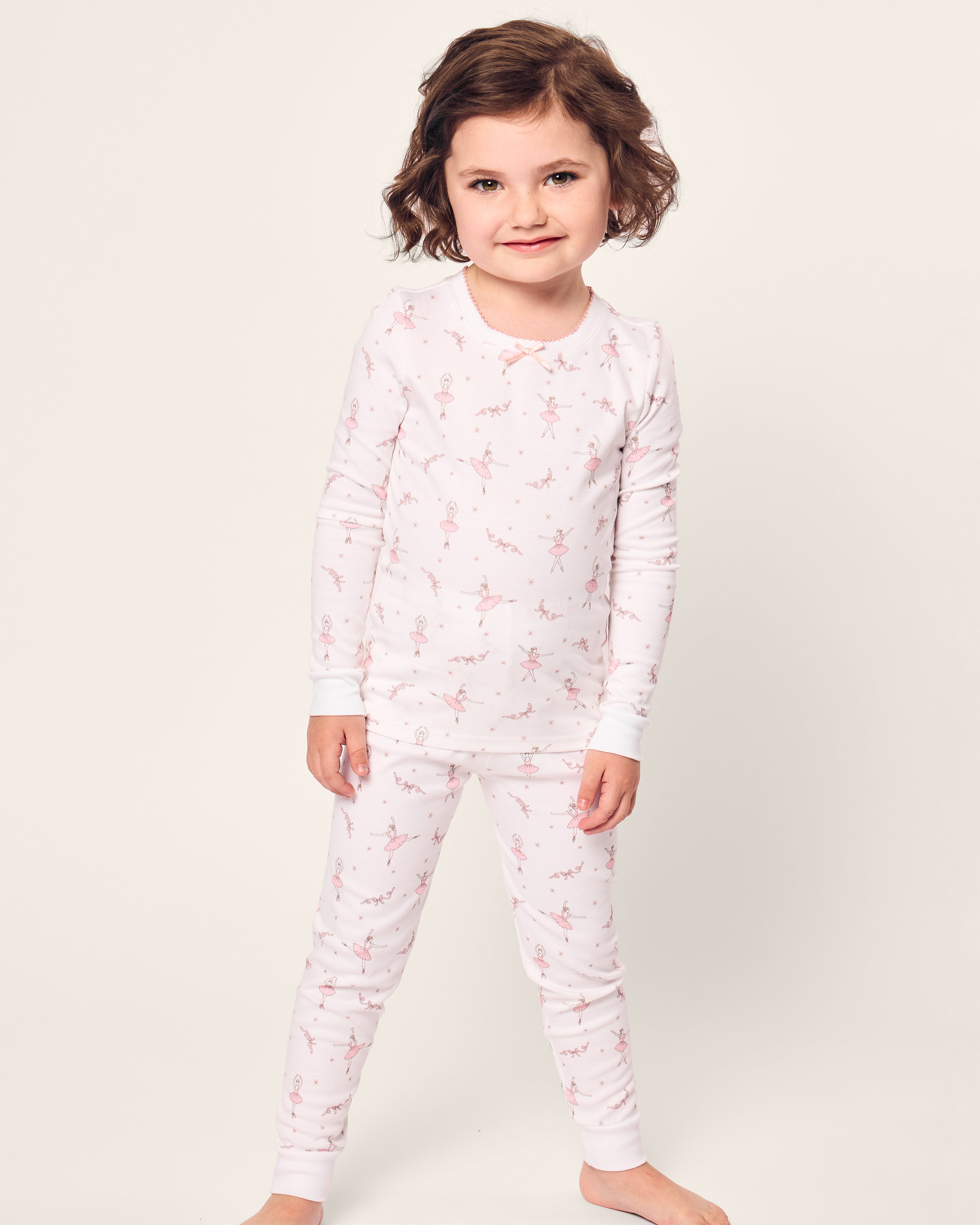 A young child with short brown hair is smiling, standing in Petite Plumes Kids Pima Snug Fit Pajama Set in Sugar Plum Fairy. The soft Pima cotton ensemble features matching light pink sleepwear and a subtle pattern, with a long sleeve top against a plain, light background.