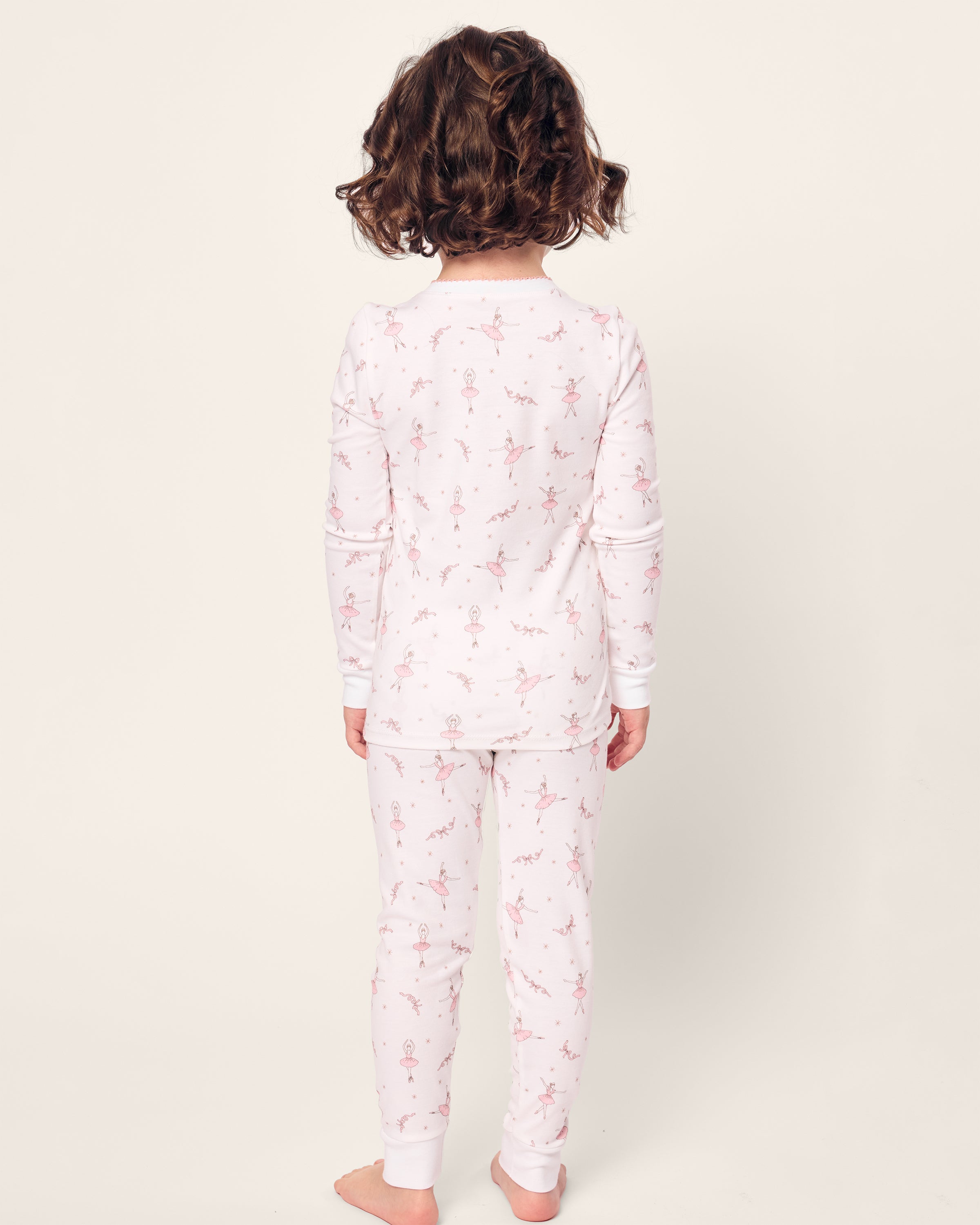 A child with curly hair wears Petite Plumes Kids Pima Snug Fit Pajama Set in Sugar Plum Fairy, featuring soft white Pima cotton and small pink floral designs. They stand barefoot in a long sleeve top, facing away on a plain white background.
