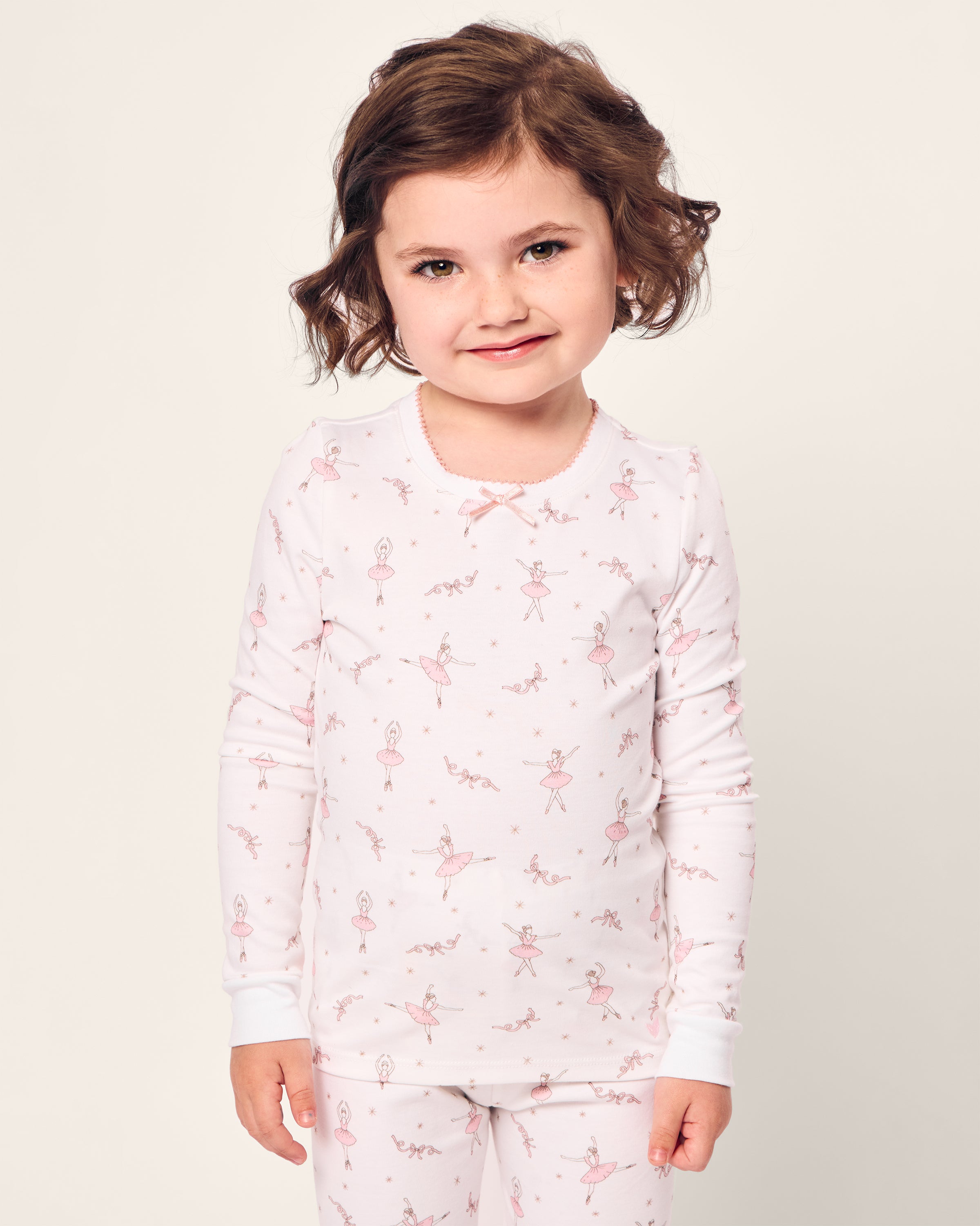 The young child beams in their Petite Plume Kids Pima Snug Fit Pajama Set in Sugar Plum Fairy, showcasing a charming ballerina and flower pattern. The long sleeve top and matching pants perfectly complement the childs short, wavy hair against a light background.
