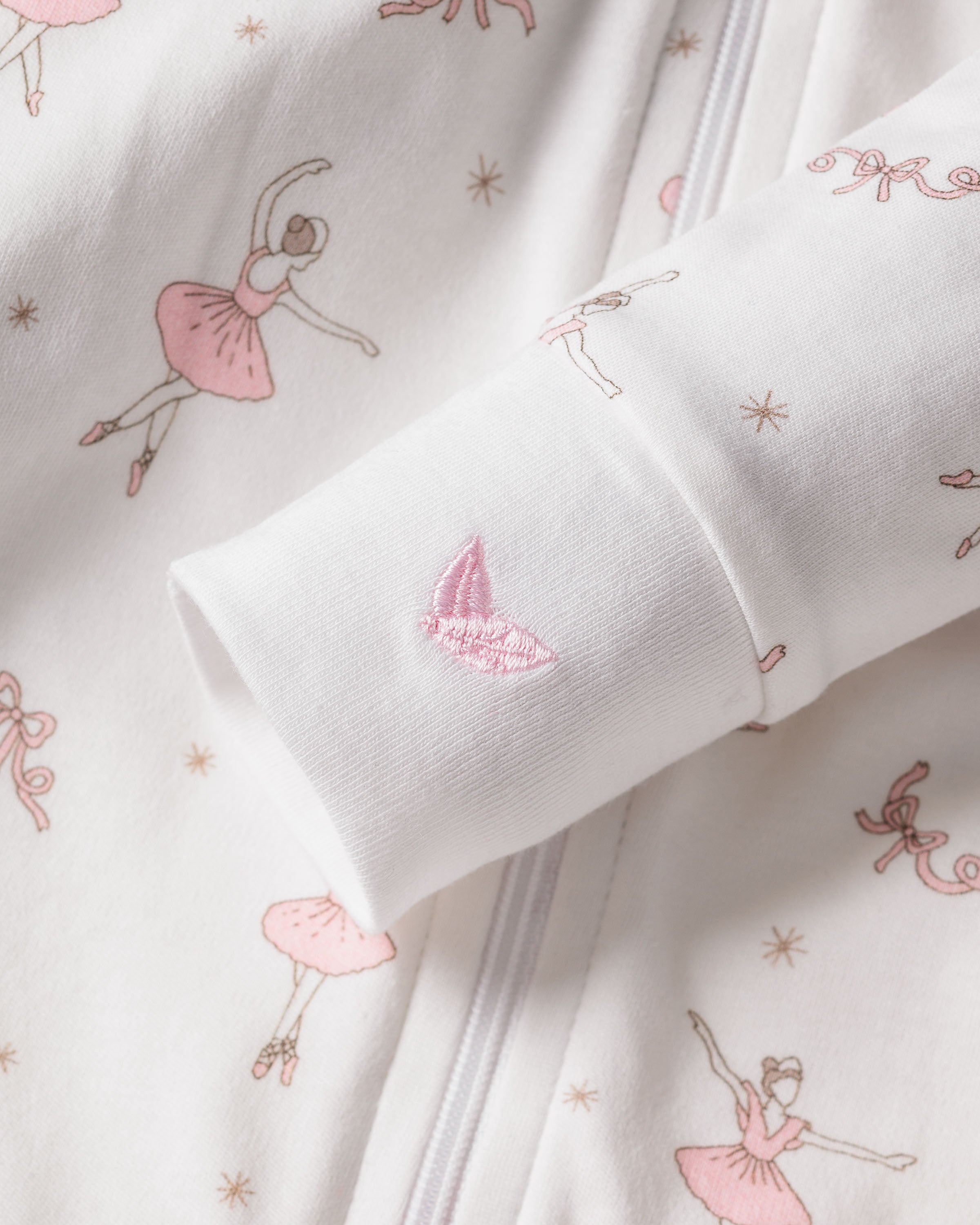 Close-up of the Babys Pima Romper in Sugar Plum Fairy from Petite Plume, showcasing white Pima cotton fabric with pink ballerina and bow patterns. The sleeve features a pink embroidered butterfly, and a visible vertical zipper suggests its sleepwear.