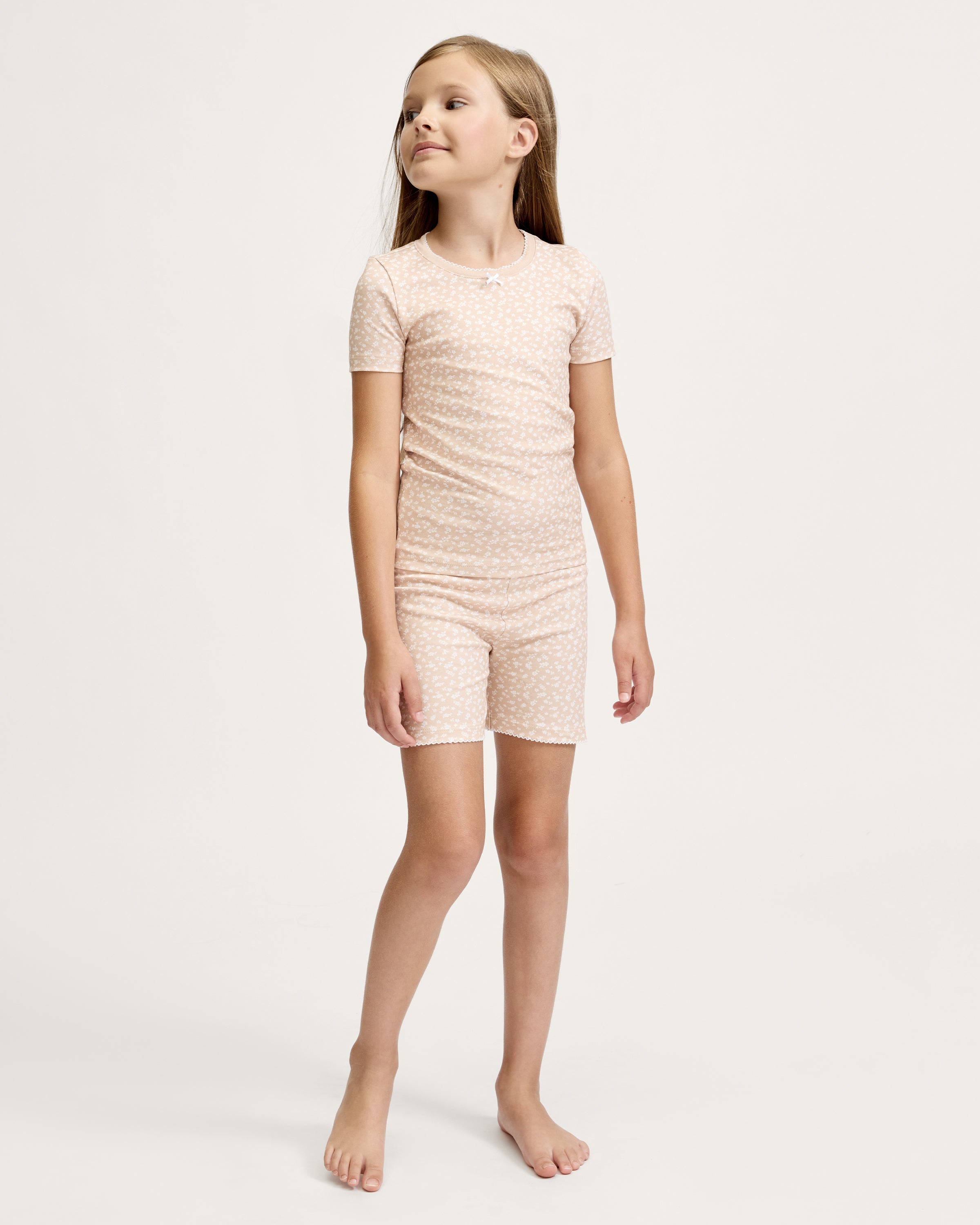 The Kid's Pima Snug Fit Short Set in Ditsy Waltz Camel is a beige, short-sleeve pajama set for kids with a subtle white floral pattern, featuring a top with a bow at the neckline and matching elastic-waist shorts.