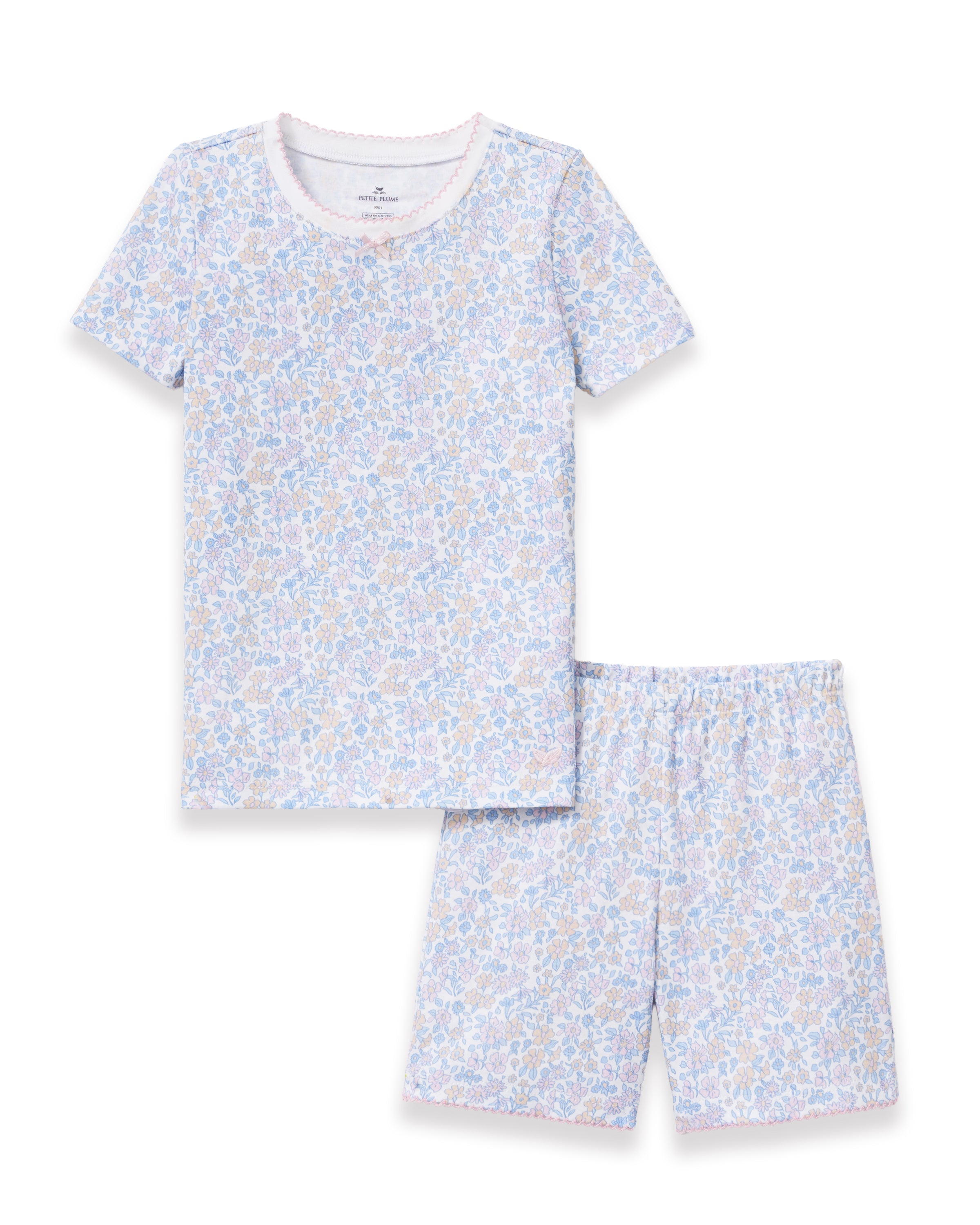 A young girl with long, wavy hair smiles with her hands on her hips, wearing the Kid's Pima Snug Fit Short Set in Fleur Blanche. She stands against a plain, light-colored background.