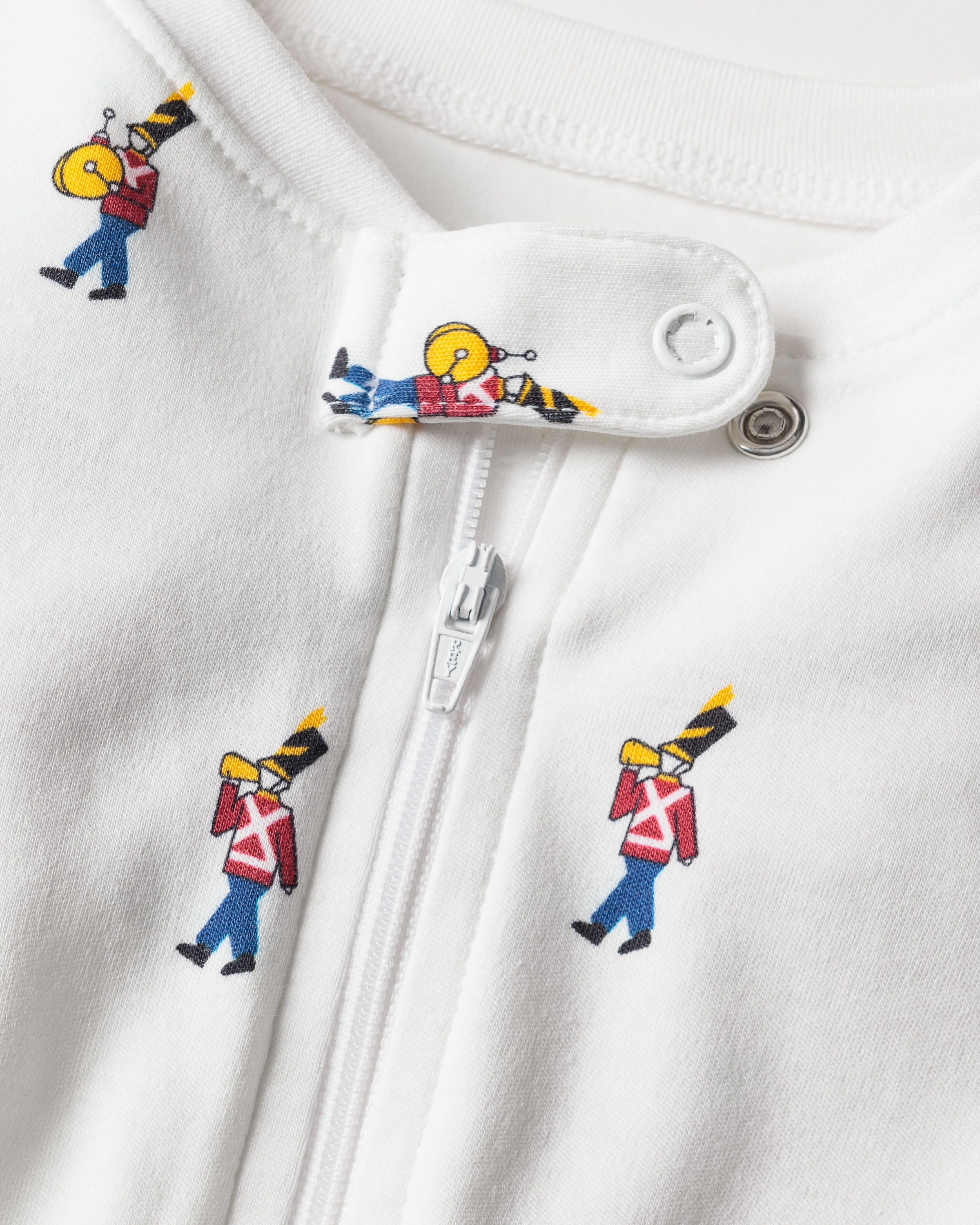 The close-up displays Petite Plumes Babys Pima Romper in Toy Soldier, crafted from soft Pima cotton. It features a zipper and snap button with a charming print of toy soldiers in red jackets, black hats, and carrying instruments repeatedly adorning the fabric.