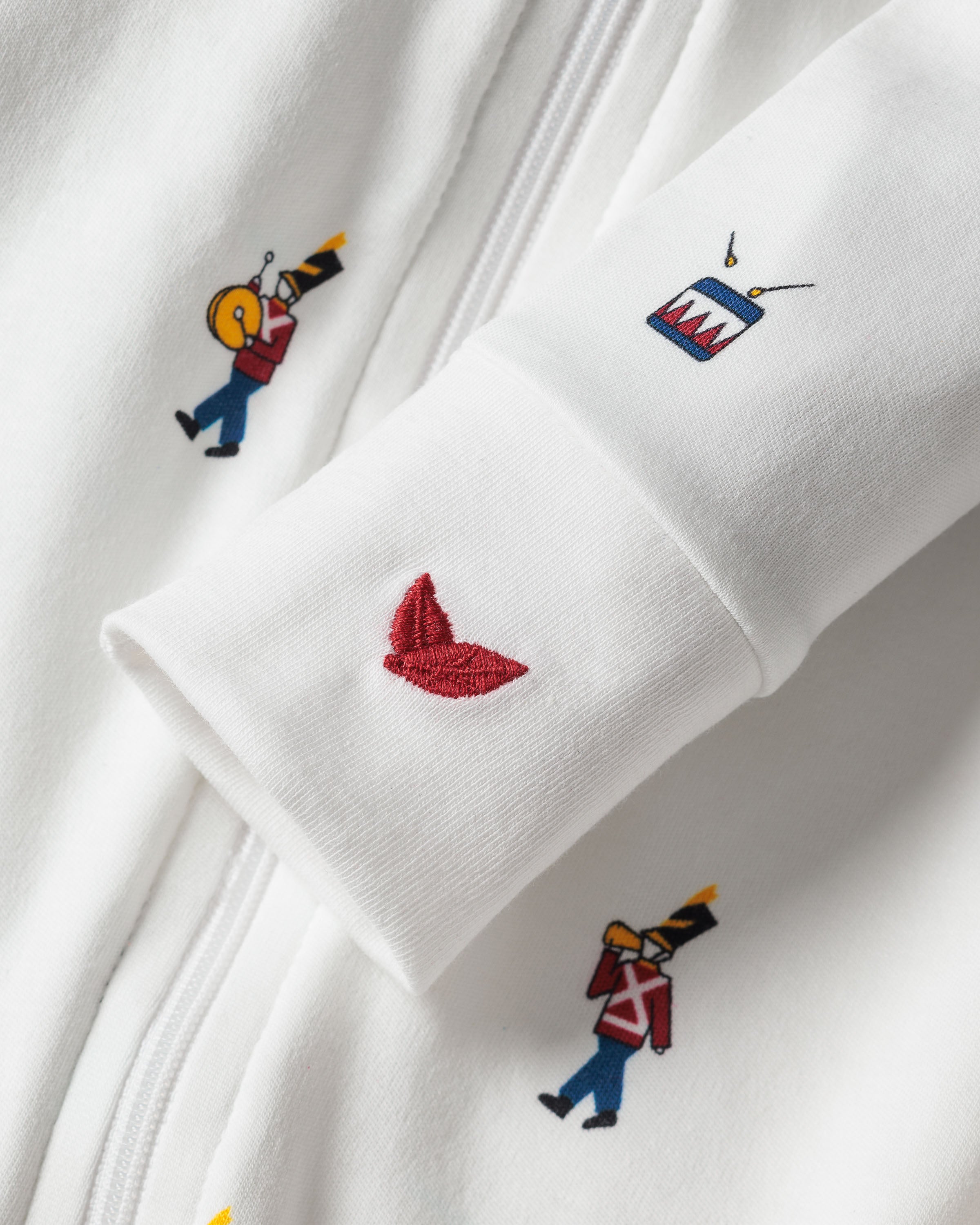 A close-up of Petite Plumes Babys Pima Romper in Toy Soldier shows its charming embroidered patterns: two red-uniformed soldiers, a red bird, and a blue box with red teeth. These designs are scattered across the fabric near the zipper.
