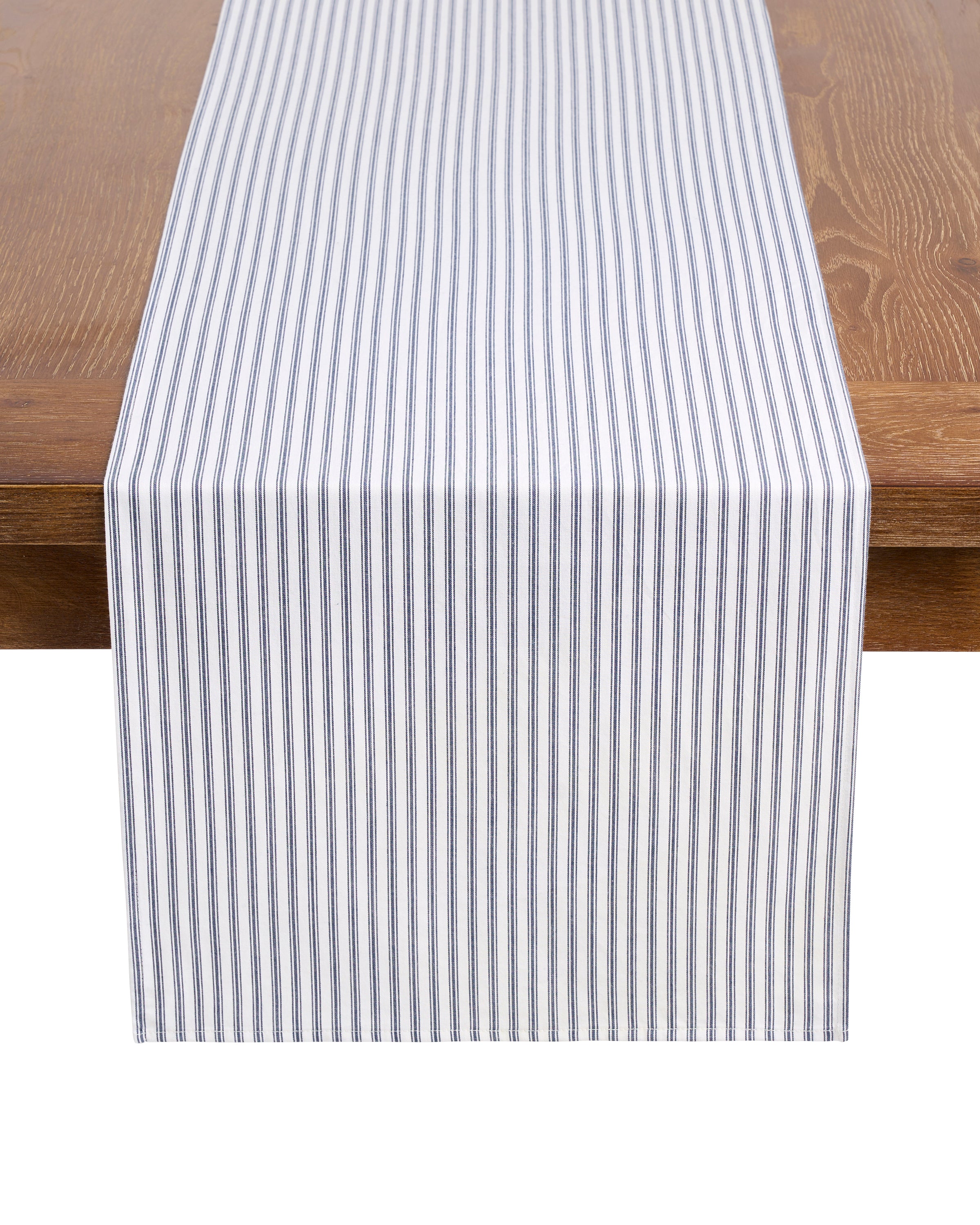 A wooden table is elegantly adorned with the Signature Twill Table Set in Navy French Ticking by Petite Plume. Its blue and white vertical stripes, crafted from 100% cotton, beautifully contrast the tabletops rich brown finish.