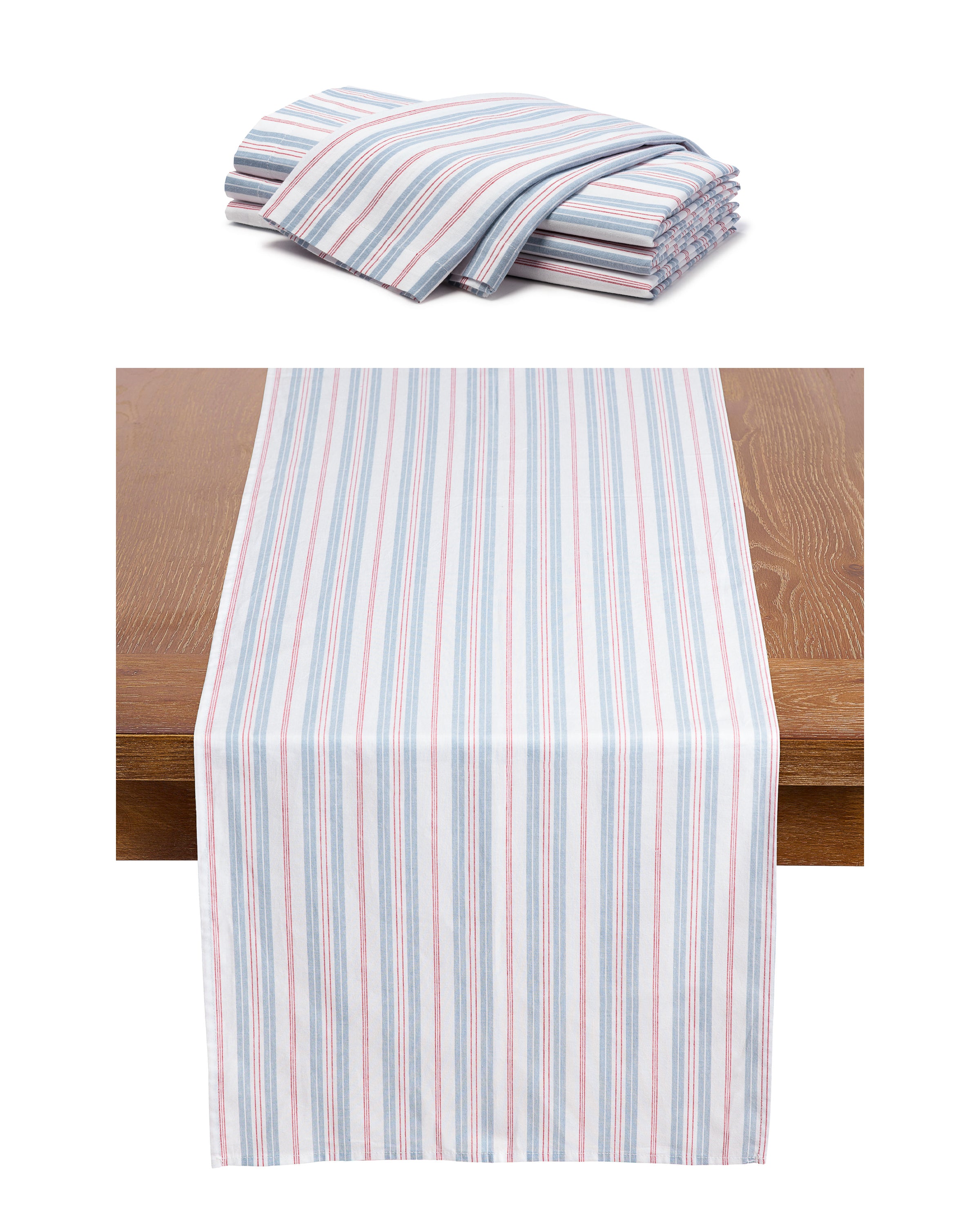 A folded pile of Signature Twill Table Runner & Napkin set in Vintage French Stripes sits above a runner on a wooden table, showcasing Petite Plumes charm. These linens boast thin red, blue, and white vertical stripes on 100% cotton.