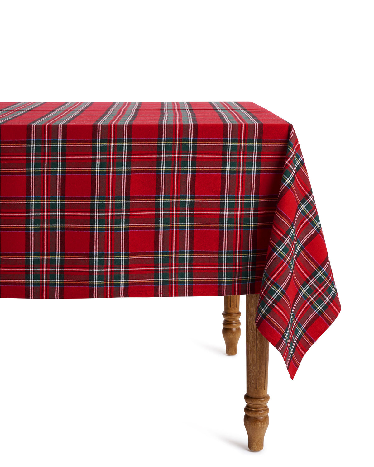 A festive tablescape features white plates on a Petite Plume Signature Twill Tablecloth in Imperial Tartan, accented with plaid napkins. Silver cutlery and crystal glasses complete the setting, while red candles and greenery add warmth beneath a softly lit Christmas tree.