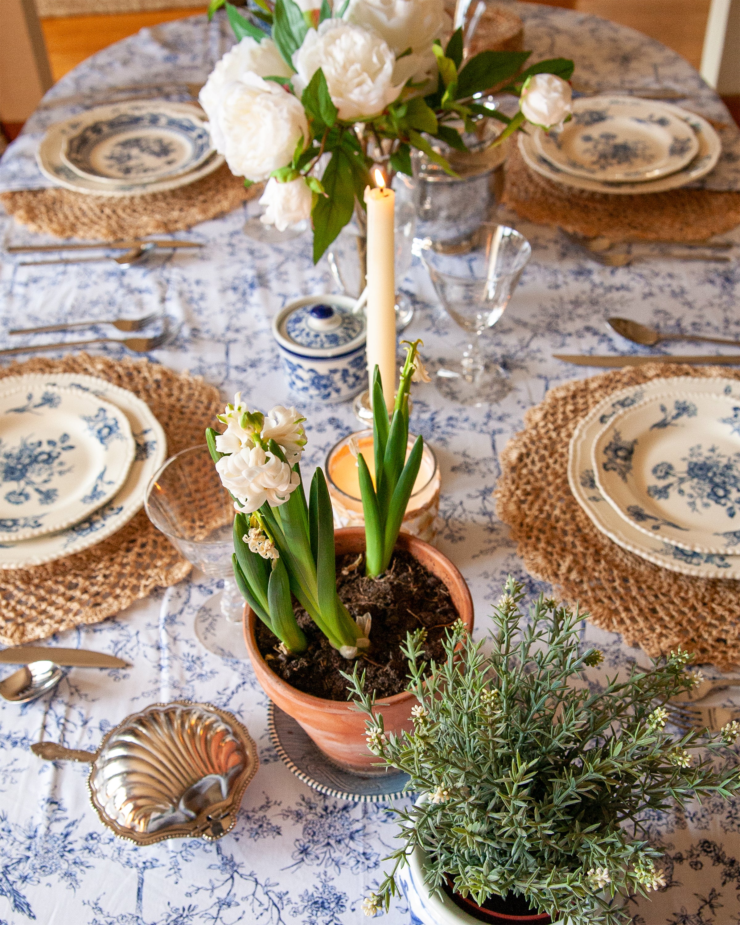 The dining table set boasts blue and white floral plates, woven placemats, and silverware, elegantly adorned with 100% cotton Petite Plume Signature Twill Tablecloth in Timeless Toile. White flowers, a candle, and potted green plants serve as the graceful centerpiece.