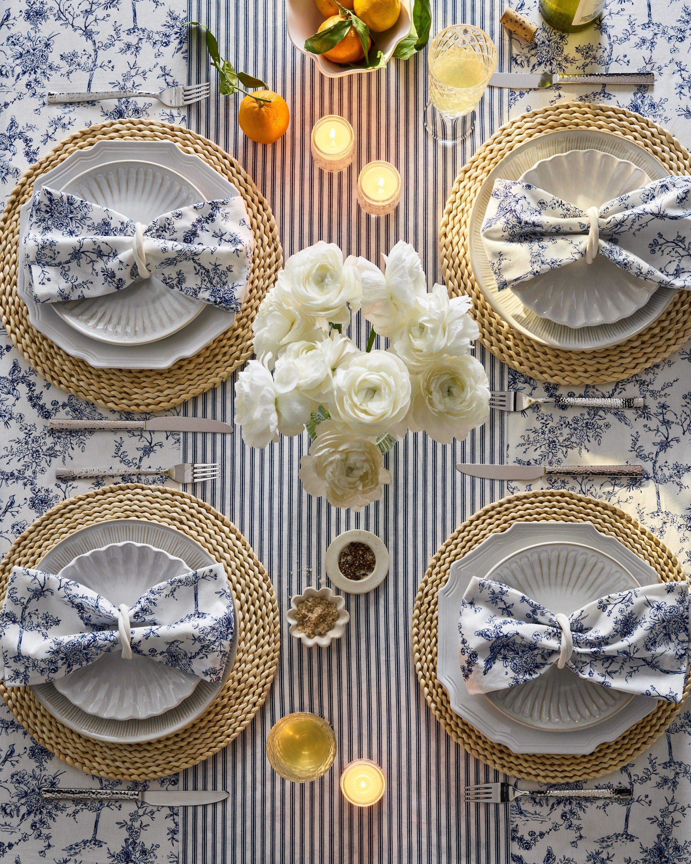 A beautifully set dining table features four white plates, blue floral napkins, and woven placemats. White flowers, lit candles, and oranges take center stage. Two glasses of white wine add elegance, while the Petite Plume Signature Twill Tablecloth in Timeless Toile completes the festive scene.