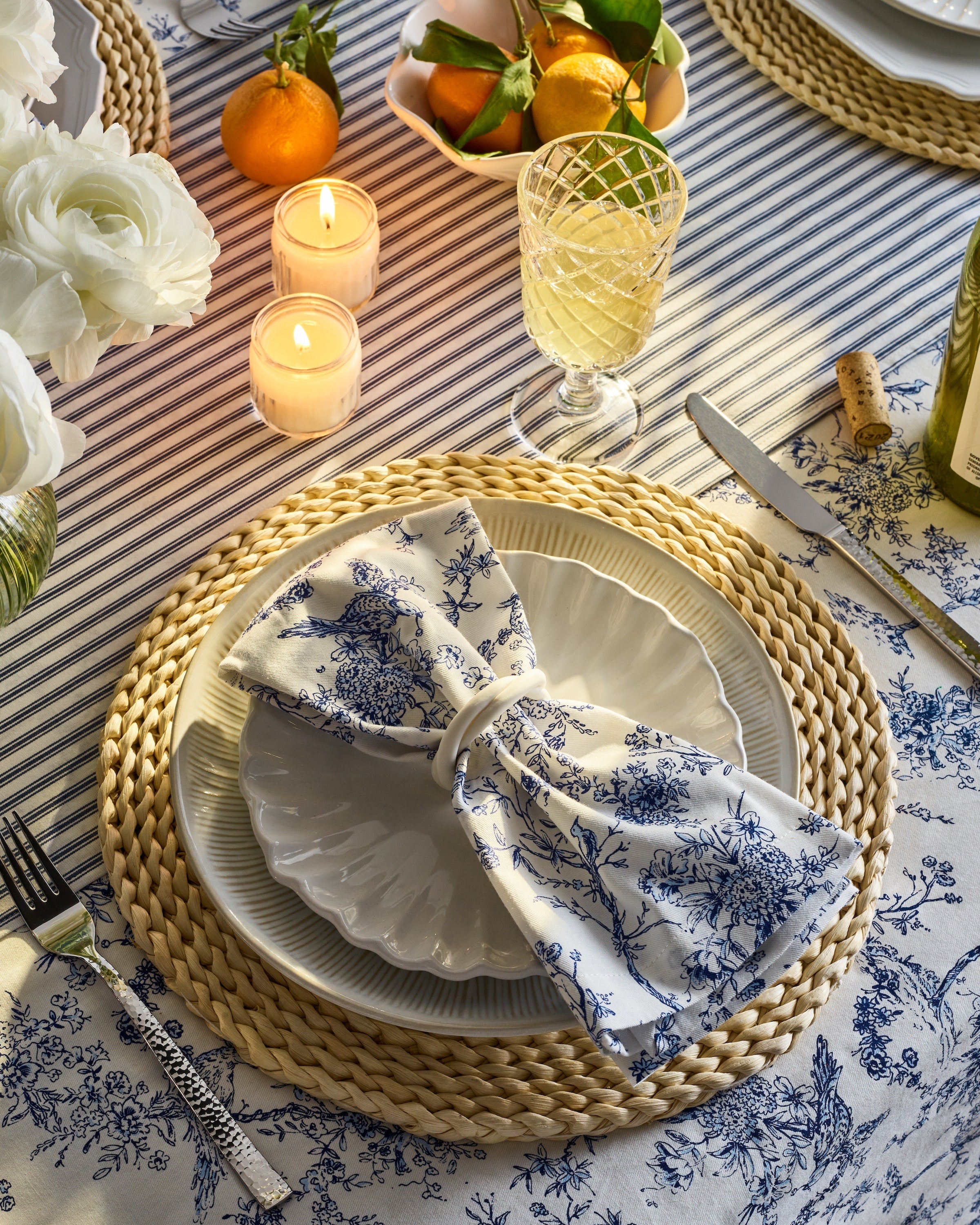 A beautifully set dining table features a blue floral napkin, crafted from 100% cotton, folded like a bow on a white plate with a wicker placemat. Surrounding it are white flowers, candles, wine, and oranges on Petite Plumes Signature Twill Tablecloth in Timeless Toile.