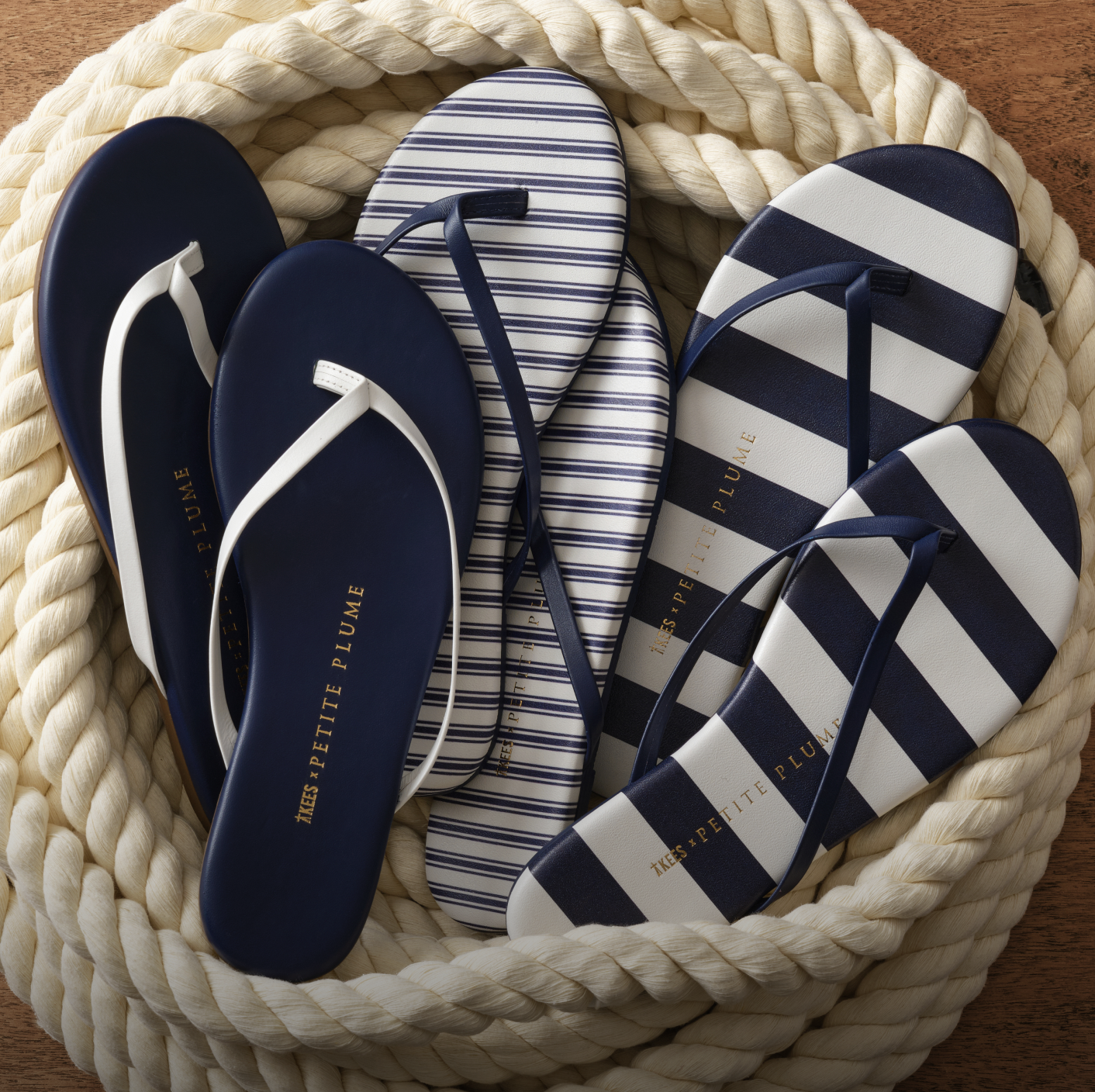 Three pairs of flip-flops with navy, white, and striped designs rest in a coiled rope basket. The flip-flops display the brand name Petite Plume in gold lettering on the soles.