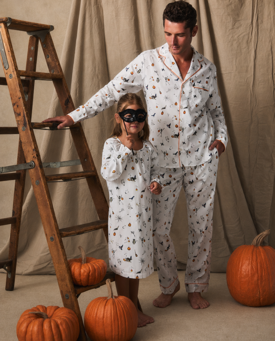 A man and a child in matching Halloween pajamas stand by a wooden ladder, surrounded by pumpkins. The child wears a black mask. Neutral brown fabric hangs in the background.