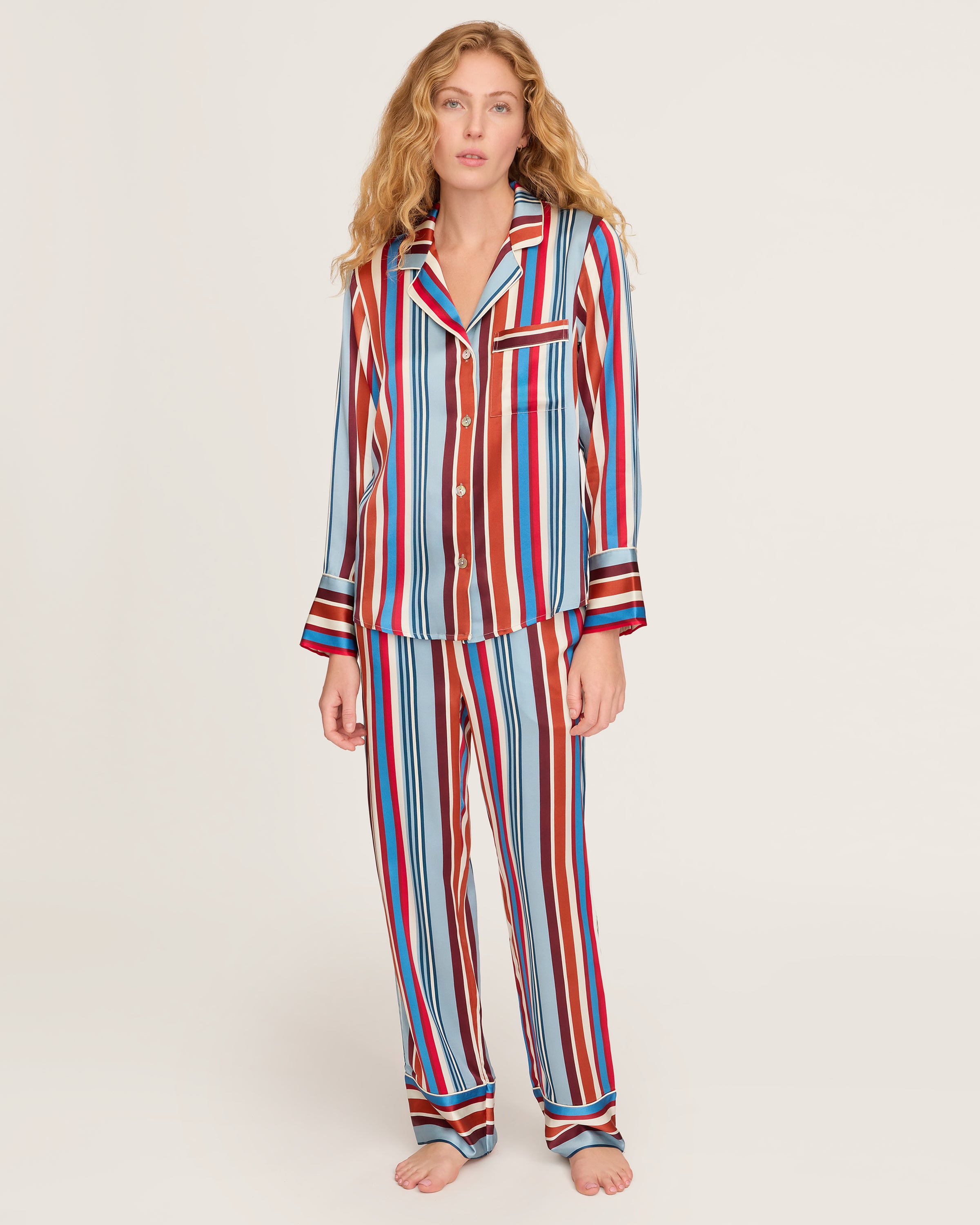 The Women's Silk Pajama Set in Discovery Stripe features long-sleeve, button-up top and matching pants with bold red, blue, brown, and white vertical stripes on smooth silk, displayed on a white background.