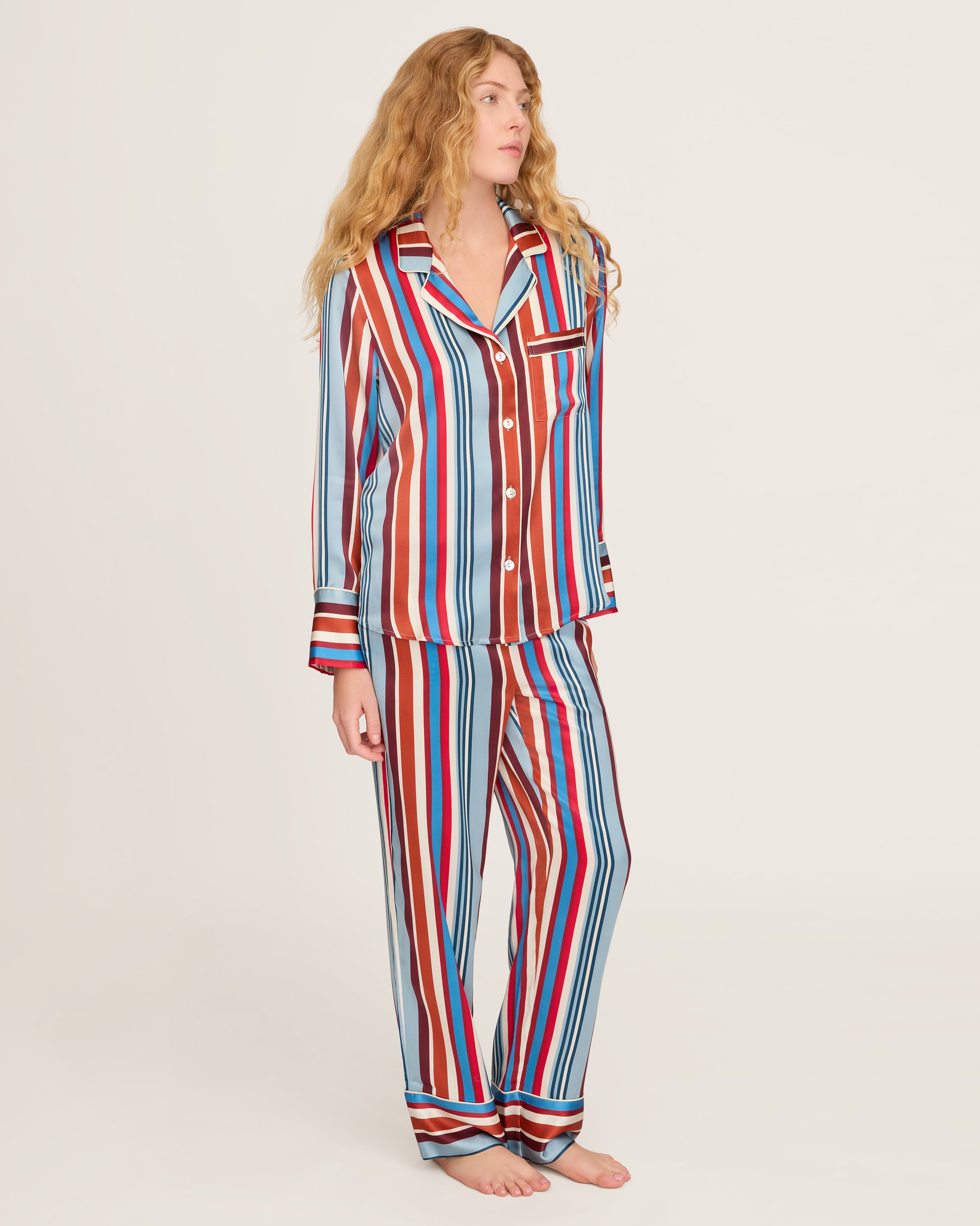 The Women's Silk Pajama Set in Discovery Stripe features long-sleeve, button-up top and matching pants with bold red, blue, brown, and white vertical stripes on smooth silk, displayed on a white background.