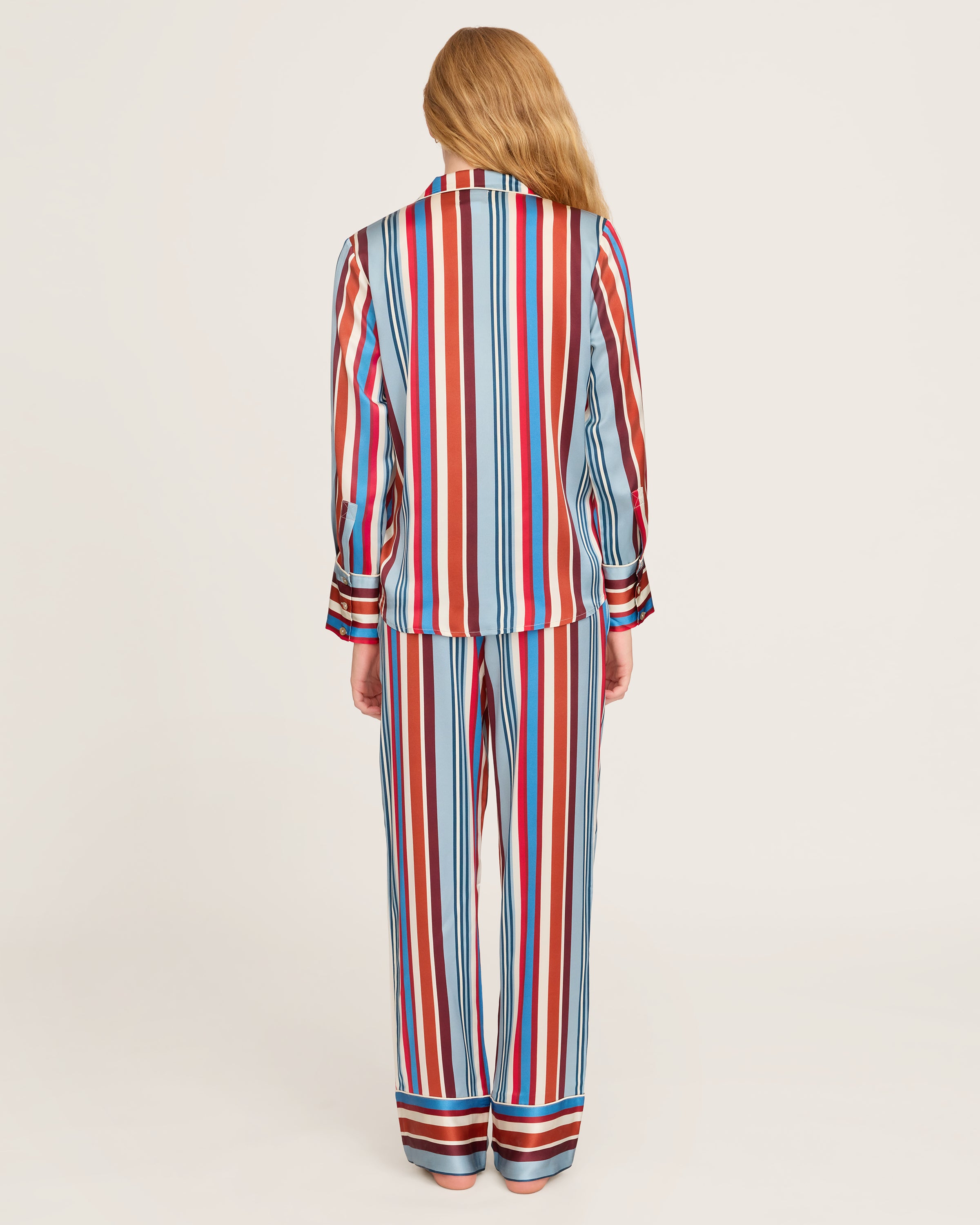 The Women's Silk Pajama Set in Discovery Stripe features long-sleeve, button-up top and matching pants with bold red, blue, brown, and white vertical stripes on smooth silk, displayed on a white background.