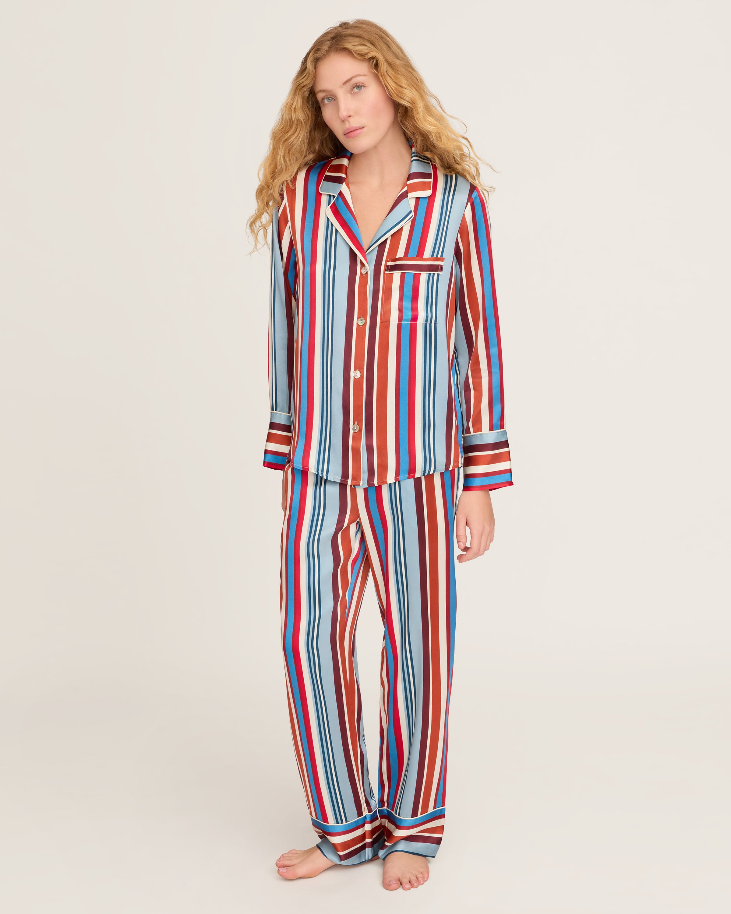 The Women's Silk Pajama Set in Discovery Stripe features long-sleeve, button-up top and matching pants with bold red, blue, brown, and white vertical stripes on smooth silk, displayed on a white background.