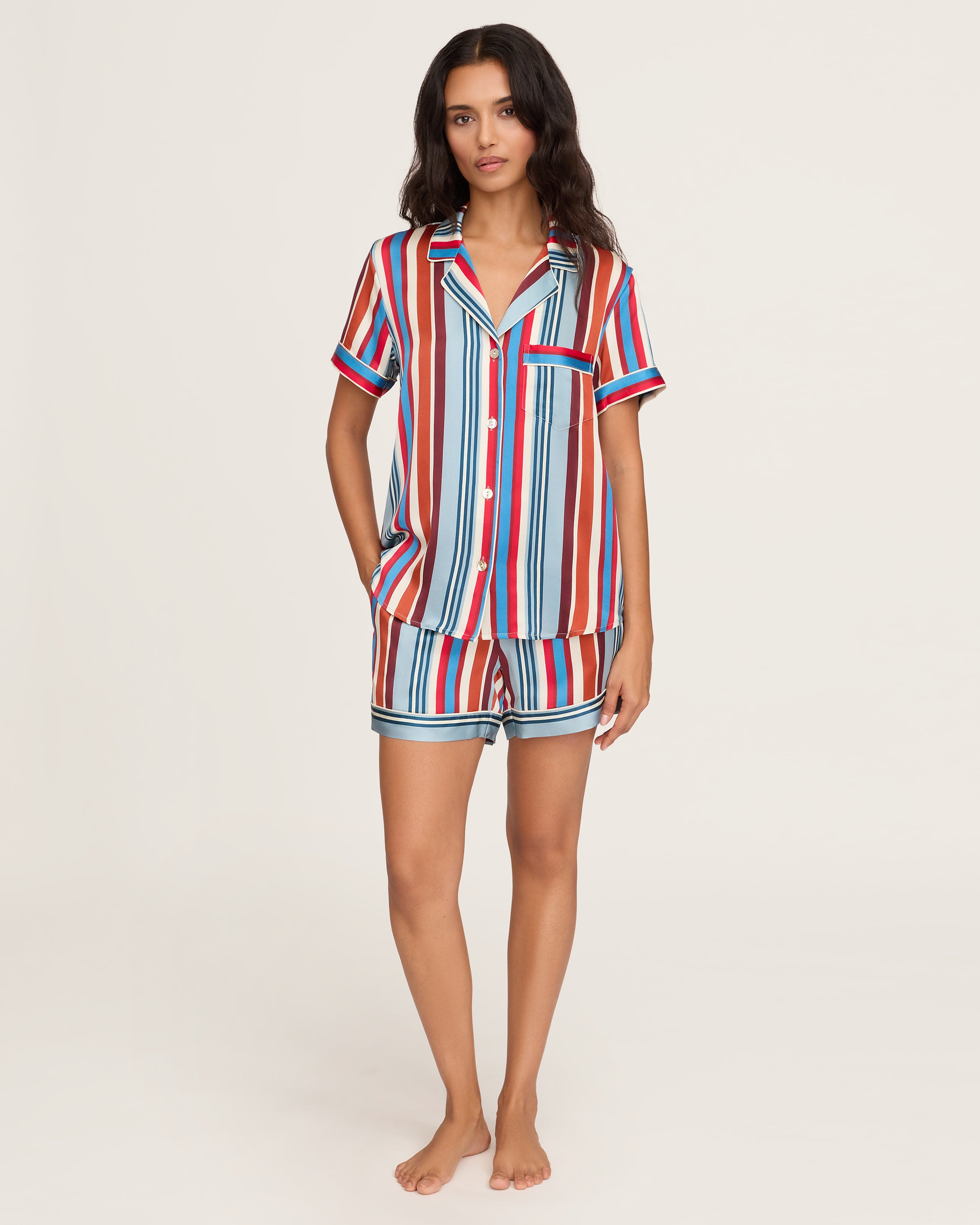 The Women's Silk Short Set in Discovery Stripe features a short-sleeved button-up shirt and matching drawstring shorts with bold vertical stripes in red, blue, brown, and white.