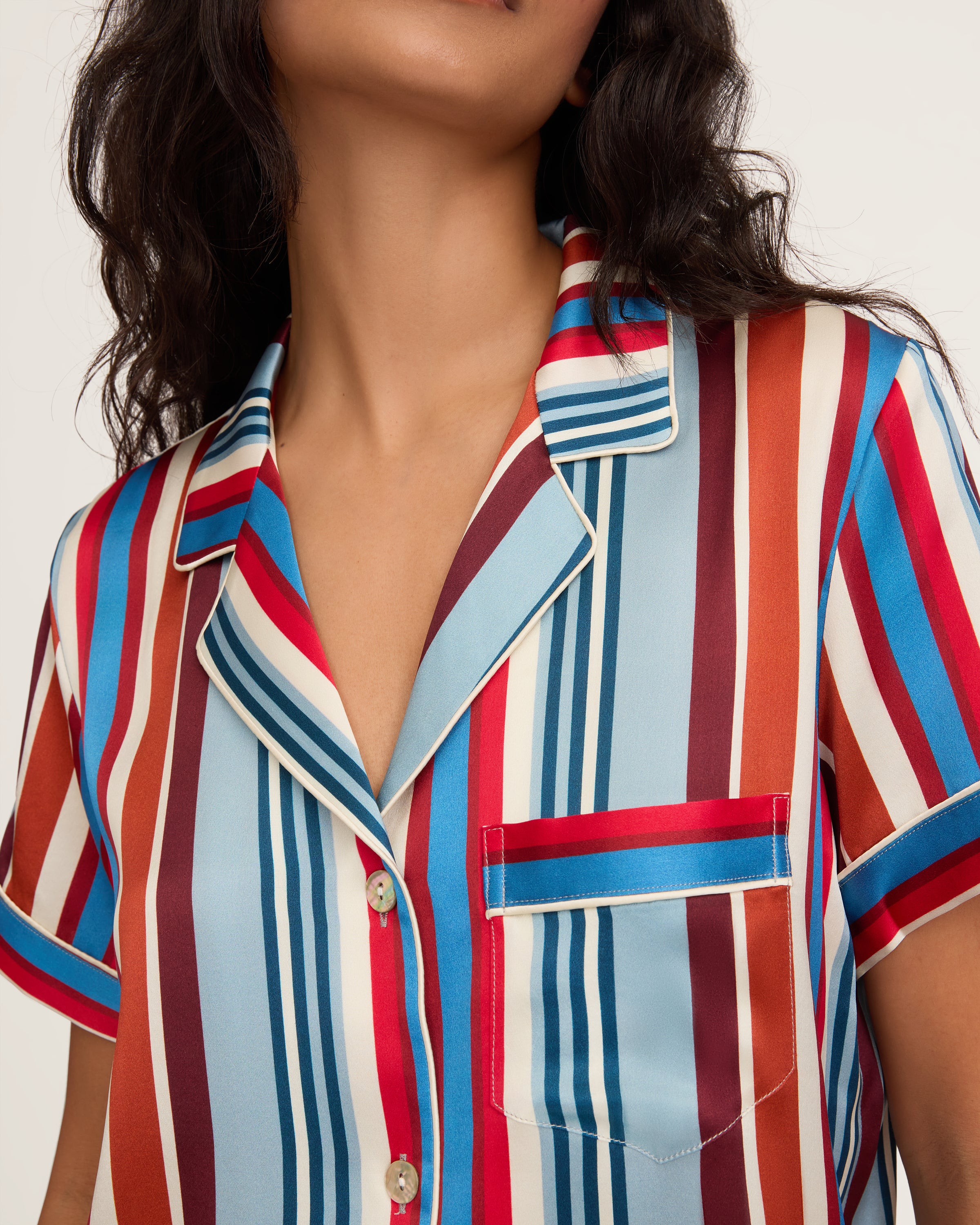 The Women's Silk Short Set in Discovery Stripe features a short-sleeved button-up shirt and matching drawstring shorts with bold vertical stripes in red, blue, brown, and white.