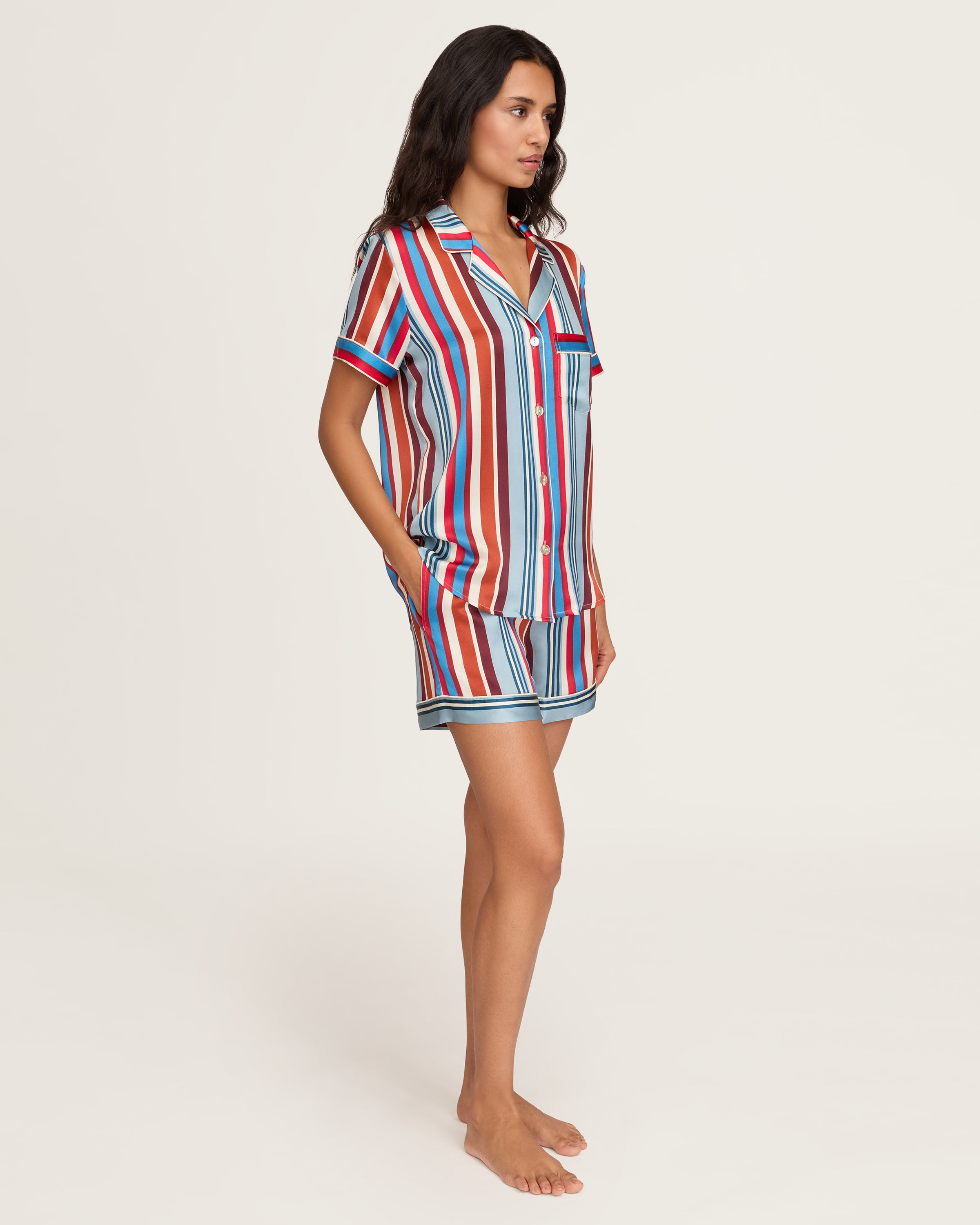 The Women's Silk Short Set in Discovery Stripe features a short-sleeved button-up shirt and matching drawstring shorts with bold vertical stripes in red, blue, brown, and white.