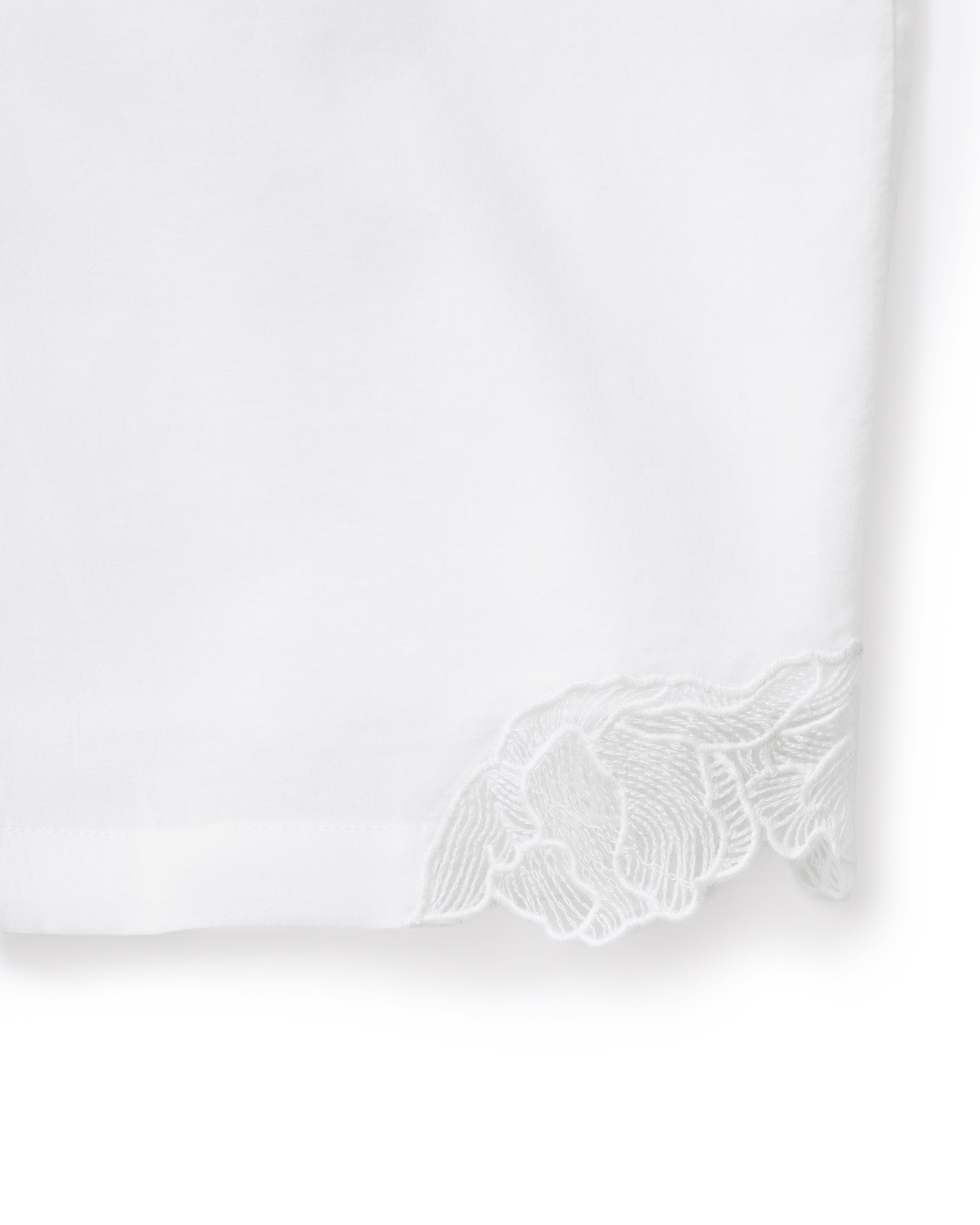A woman with wavy hair stands barefoot on a light background, wearing the Women's Voile Fleur Cami Pant Set in White—featuring a sleeveless top and pants with lace trim. Her arms are behind her back.
