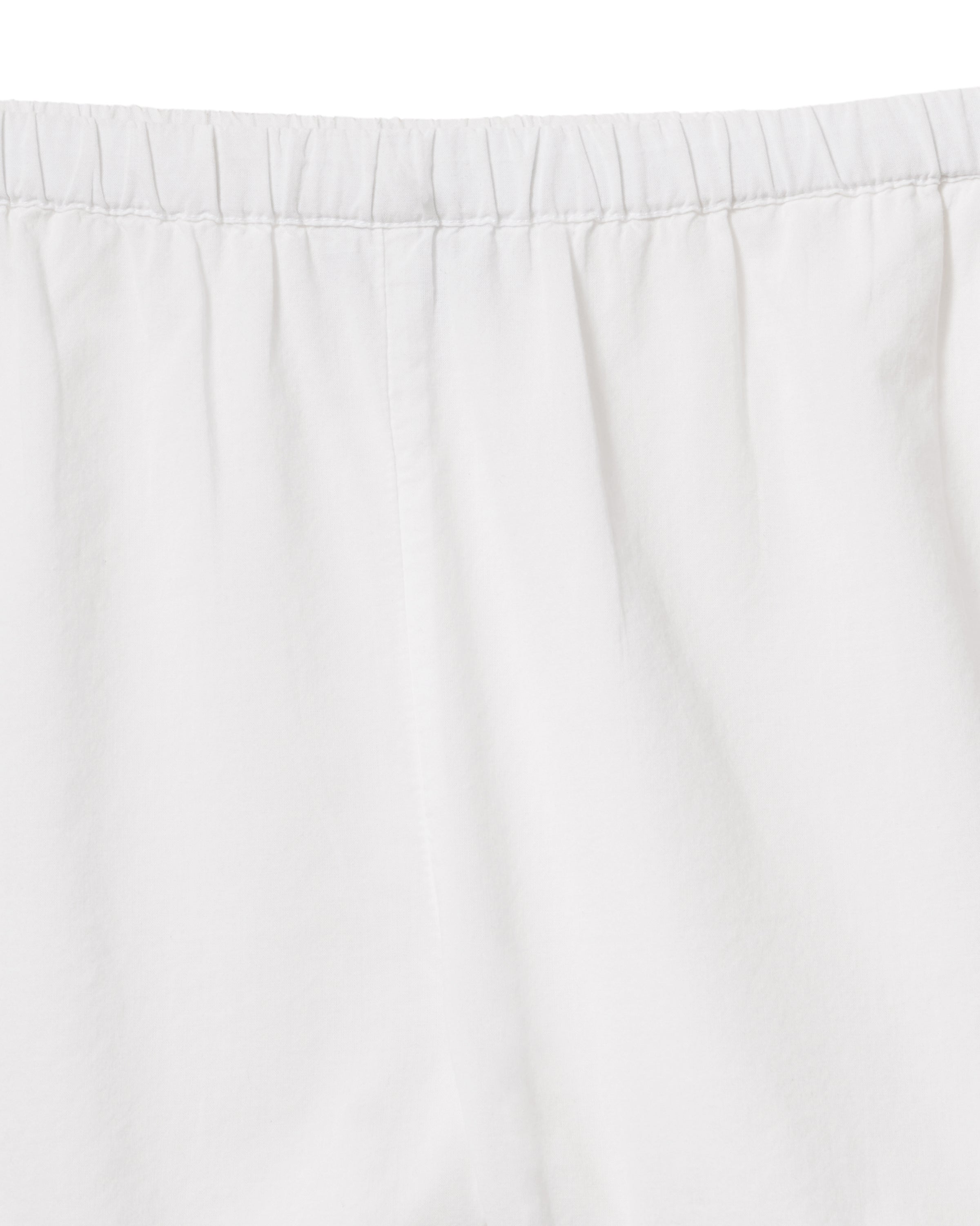 A woman with wavy hair stands barefoot on a light background, wearing the Women's Voile Fleur Cami Pant Set in White—featuring a sleeveless top and pants with lace trim. Her arms are behind her back.