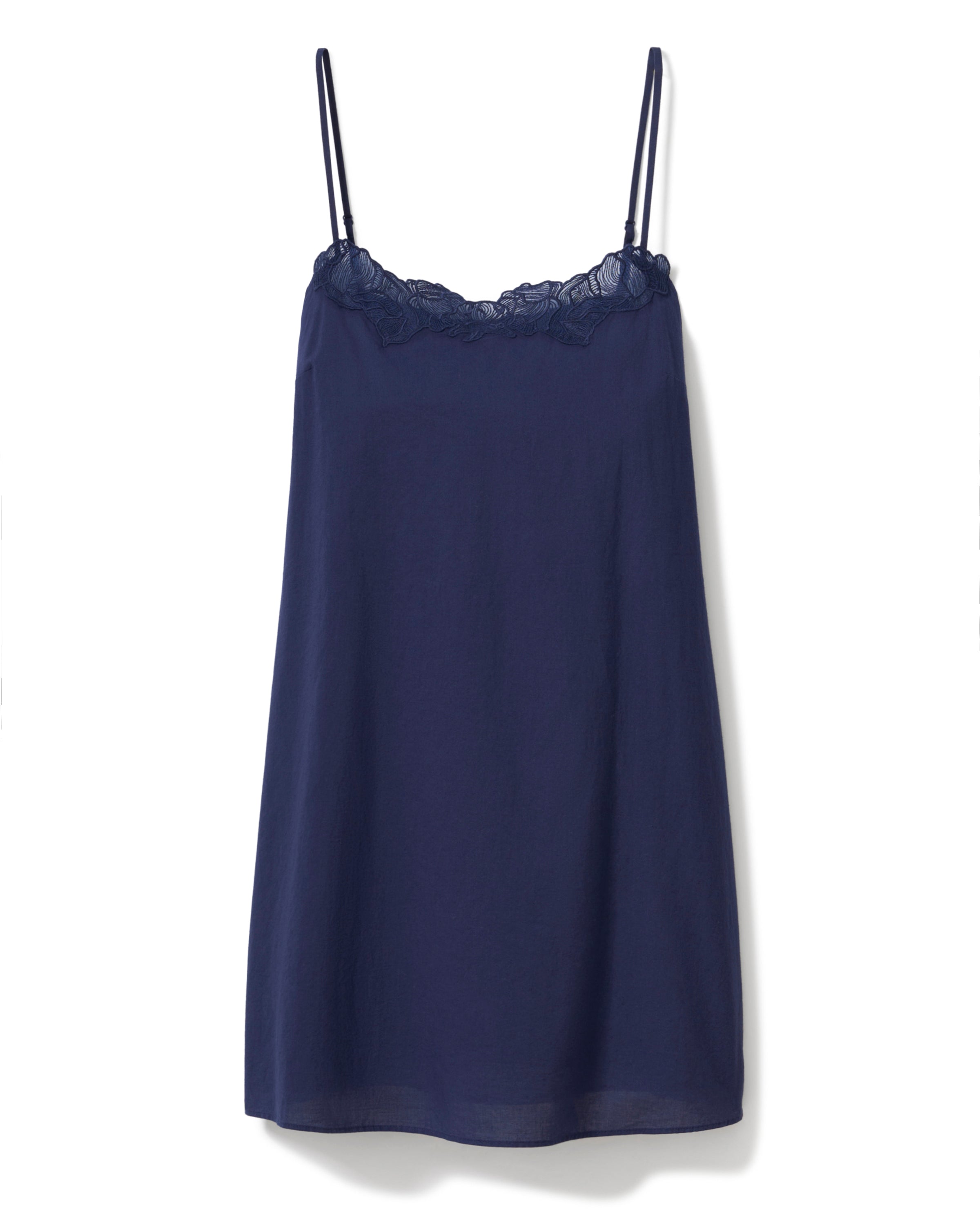 A woman with long curly hair wears the Women's Voile Fleur Slip in Navy, a sleeveless nightdress with lace trim at the neckline. She stands barefoot against a plain, light background, facing forward and smiling softly.