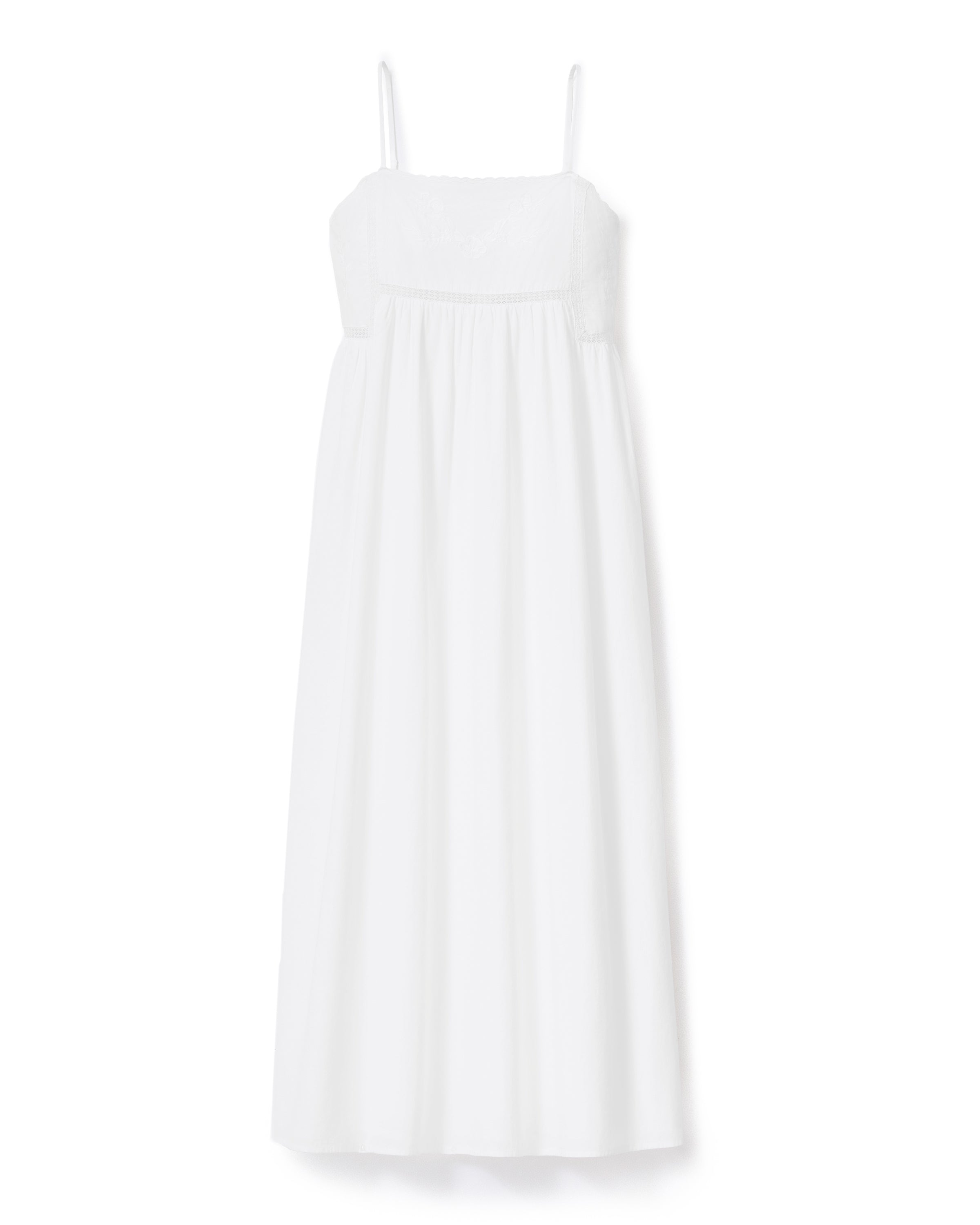 A woman with wavy hair stands barefoot and smiling to the side, wearing the Women's Voile Luna Gown in White—a long, sleeveless dress with thin straps—against a plain, light-colored background.