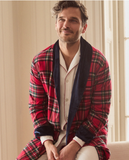 A man with wavy brown hair smiles softly while sitting indoors, wearing a red plaid robe over white pajamas with red piping. Light streams in from tall windows behind him.