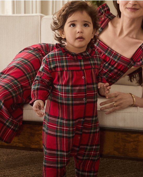 A toddler in red plaid pajamas stands in front of a woman, also wearing red plaid, who is lounging on a sofa. Both have dark hair and are indoors, creating a cozy, coordinated look.