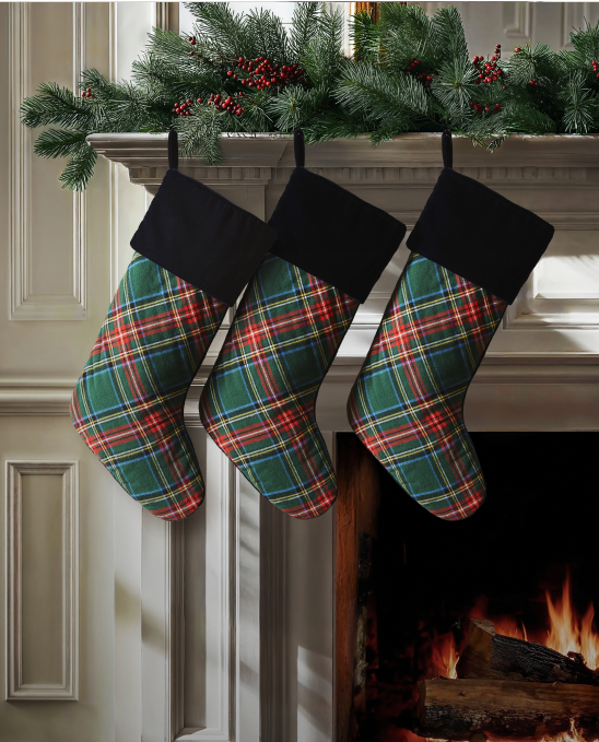 Three plaid Christmas stockings with black cuffs hang from a mantle decorated with pine branches and red berries above a lit fireplace in a cozy living room.