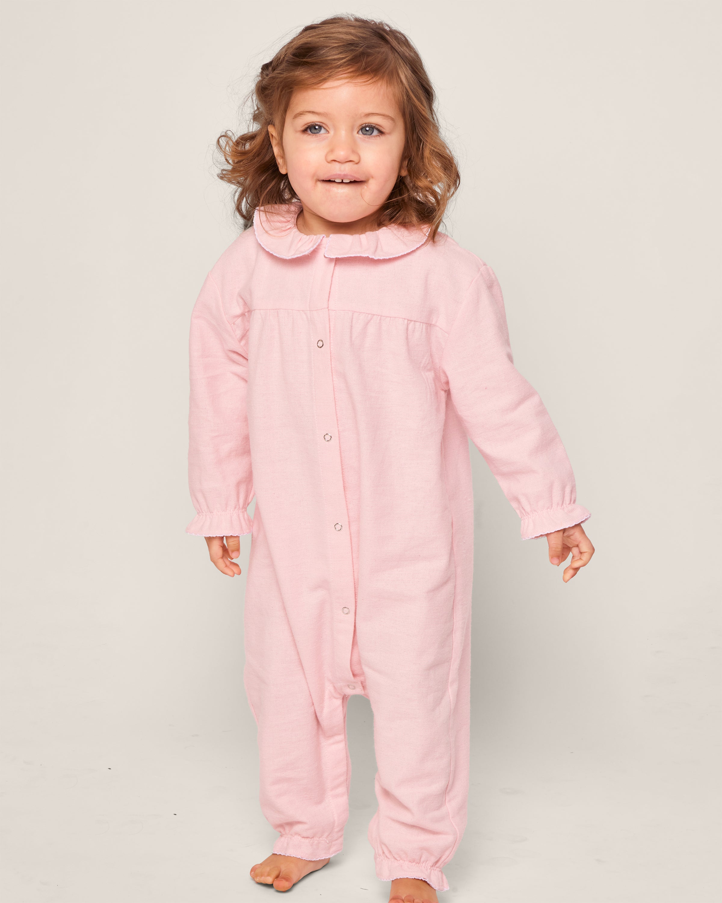 A young child with wavy brown hair is wearing Petite Plumes Babys Flannel Madeline Romper in Pink, featuring a Peter Pan collar and made of chemical-free flame-retardant material. They stand barefoot against a plain background.