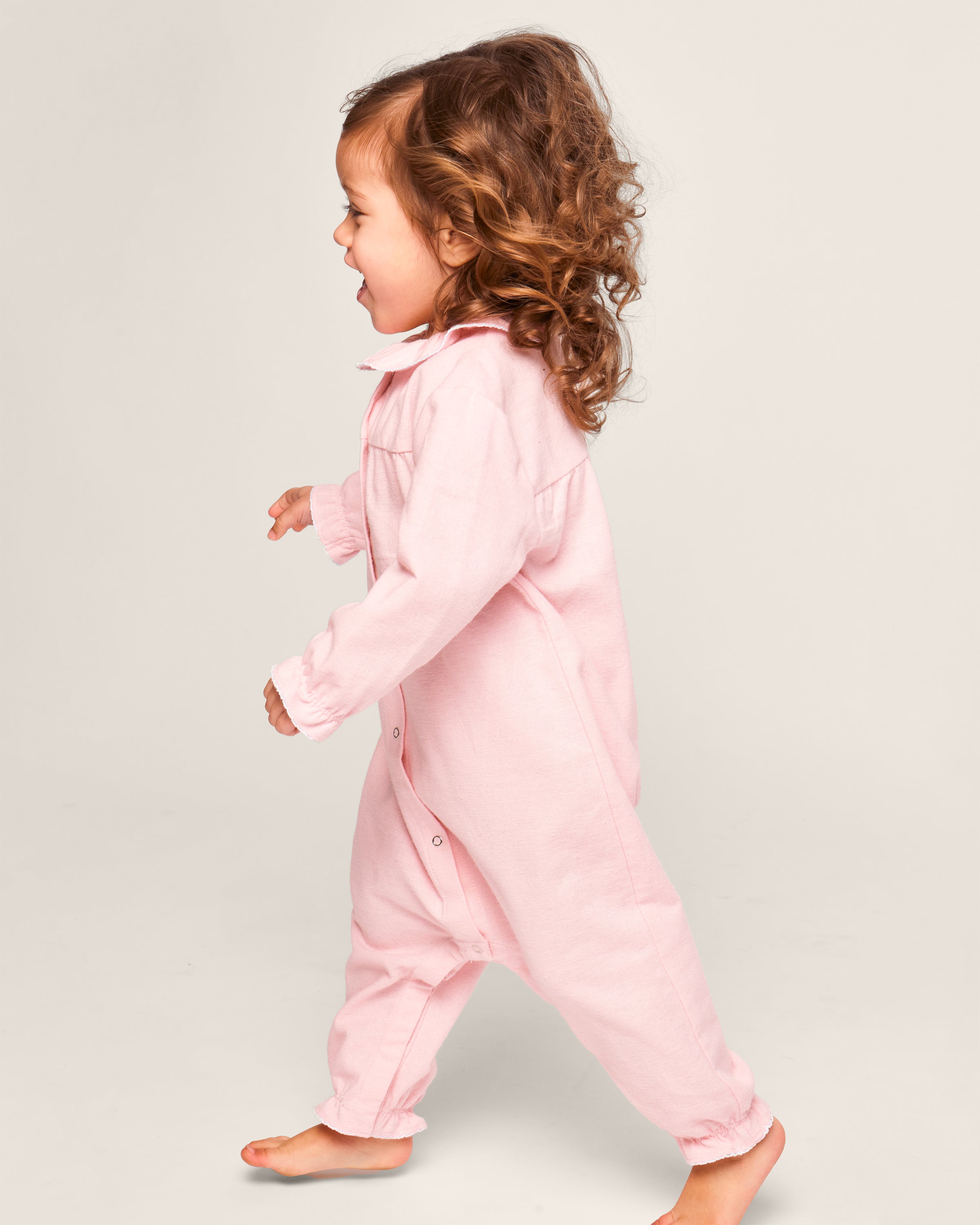 A curly-haired toddler joyfully walks in Petite Plumes Babys Flannel Madeline Romper in Pink, a brushed cotton onesie with front buttons. Theyre barefoot against a plain, neutral background.