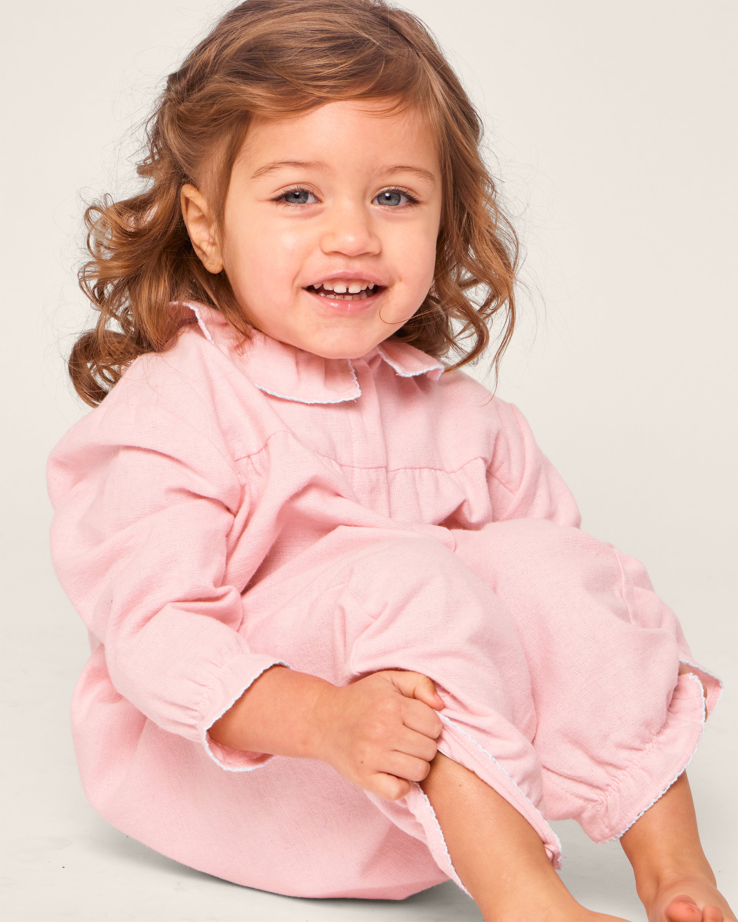 A curly-haired child smiles while sitting in Petite Plumes Babys Flannel Madeline Romper in Pink, featuring a long-sleeved jumpsuit with a ruffled collar. Made from brushed cotton flannel, it offers cozy comfort and chemical-free flame-retardant protection.