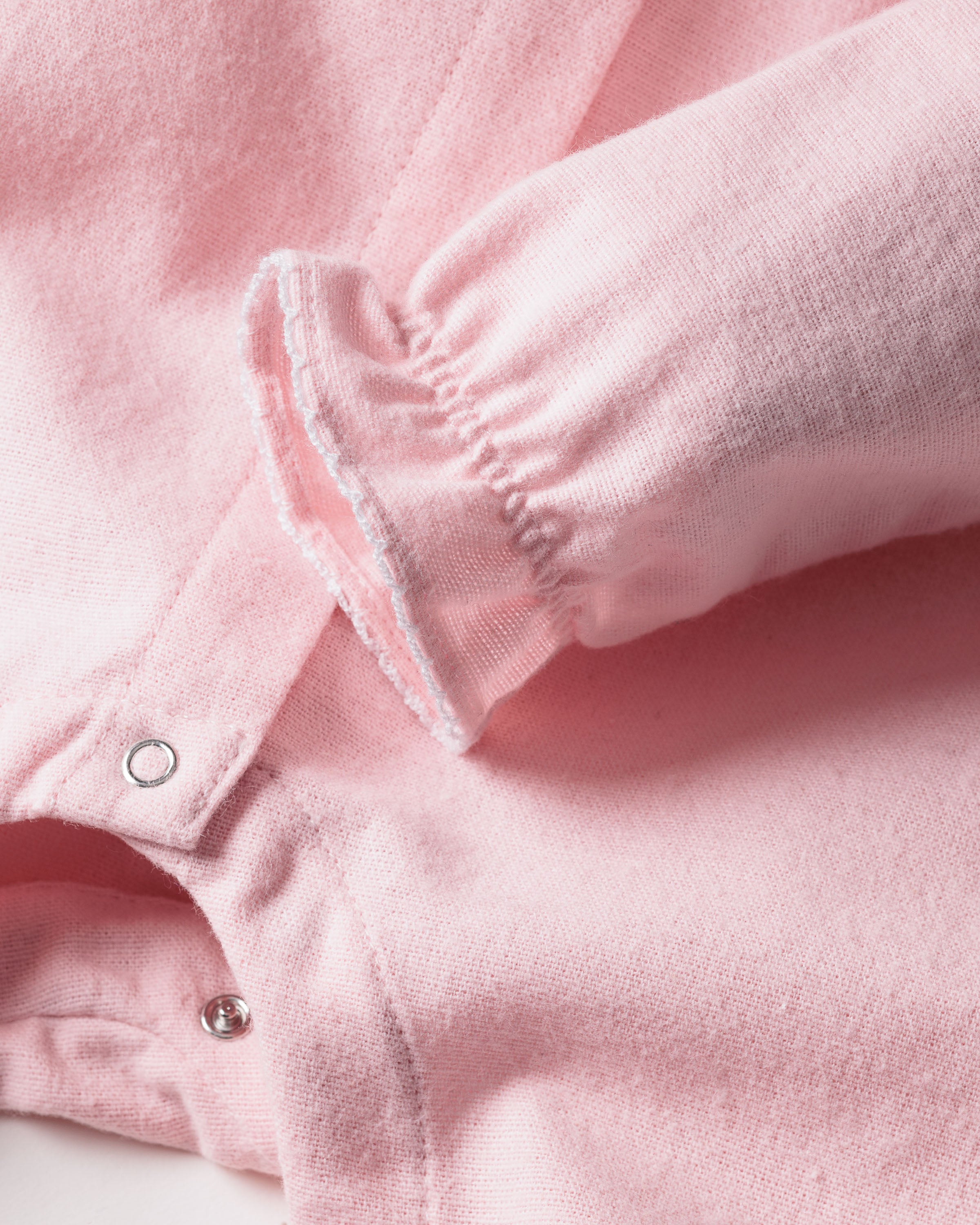 A close-up of Petite Plumes Babys Flannel Madeline Romper in pastel pink shows a brushed cotton flannel featuring long sleeves with elastic ruffle details and silver snap buttons on the front placket. The texture is soft and fleece-like, perfect for childrens sleepwear.