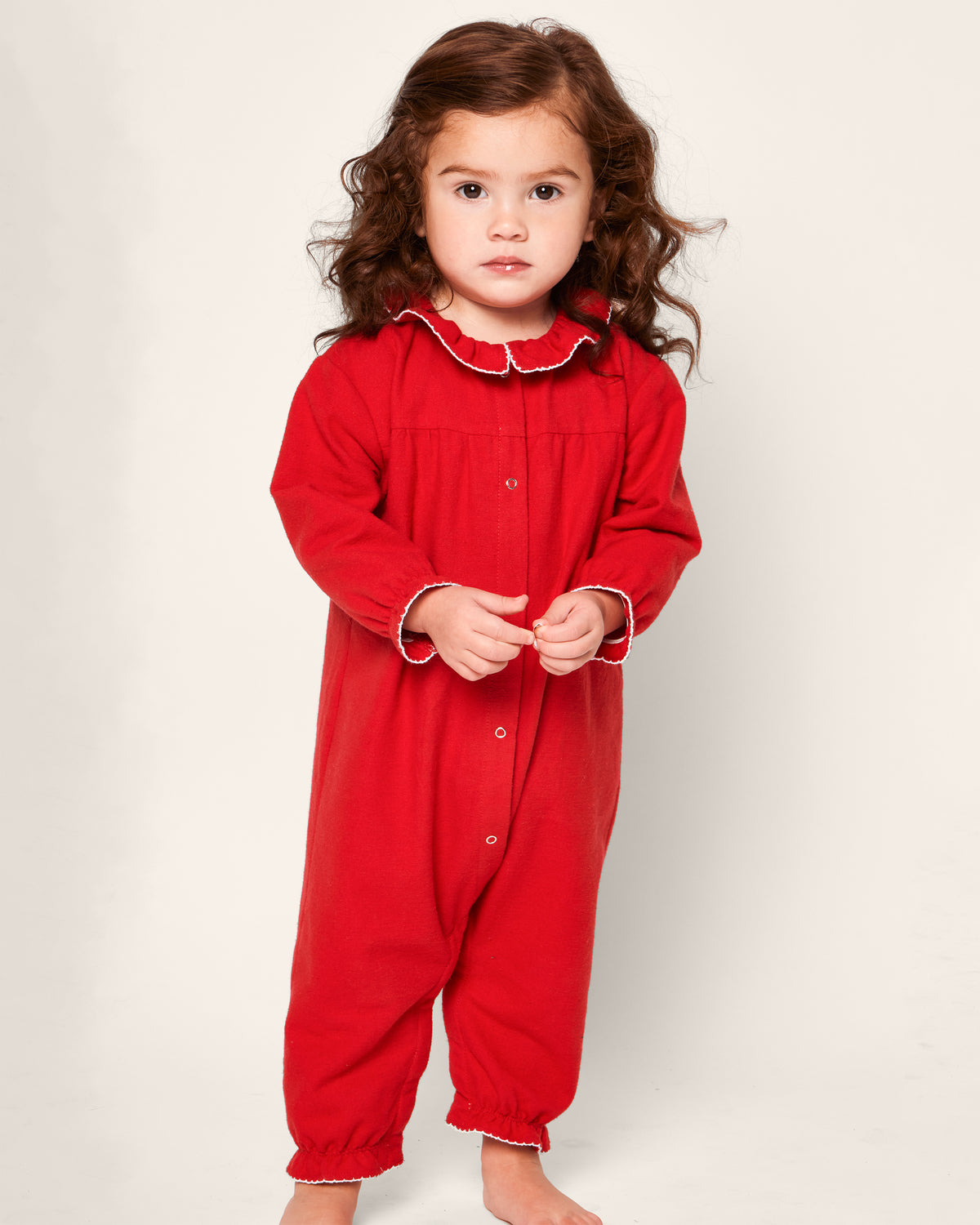A young child with curly brown hair stands barefoot in the Babys Flannel Madeline Romper in Red by Petite Plume, featuring button details and a ruffled collar, set against a plain background.