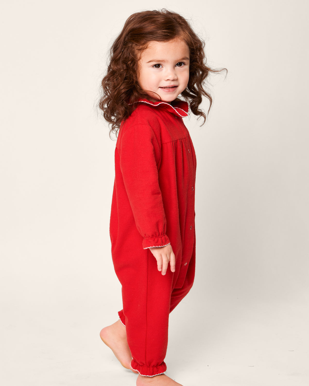A smiling young child with long brown hair is barefoot in a Petite Plume Babys Flannel Madeline Romper in red, featuring white trim. Crafted from chemical-free materials, the outfit offers comfort and safety against a pristine white backdrop.