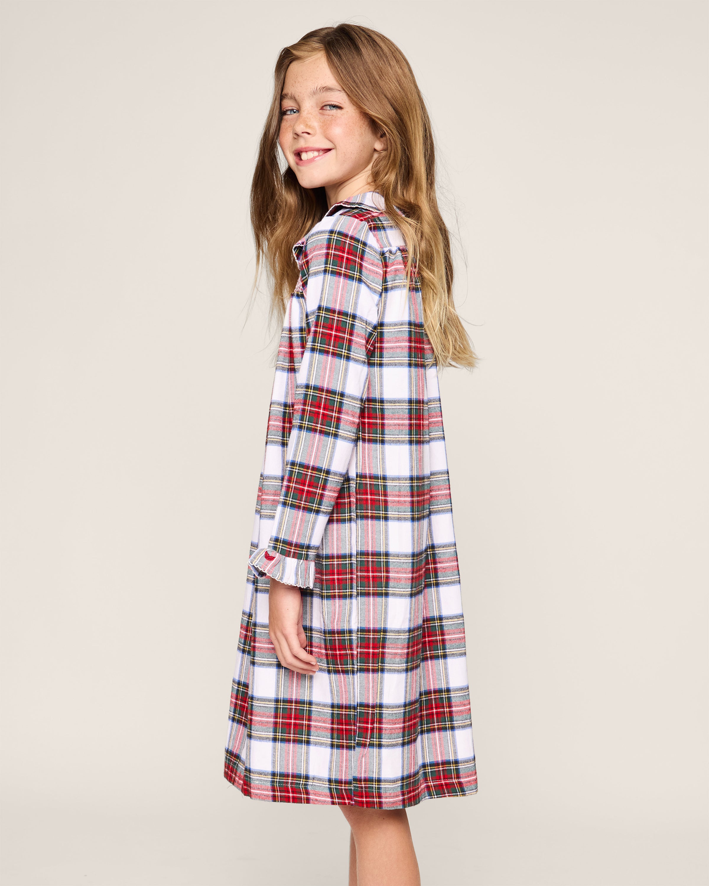 A young girl beams in a Petite Plume Girls Brushed Cotton Eloise Nightgown, showcasing red, white, and green Balmoral Tartan. Her long light brown hair cascades over her shoulders against a plain background.