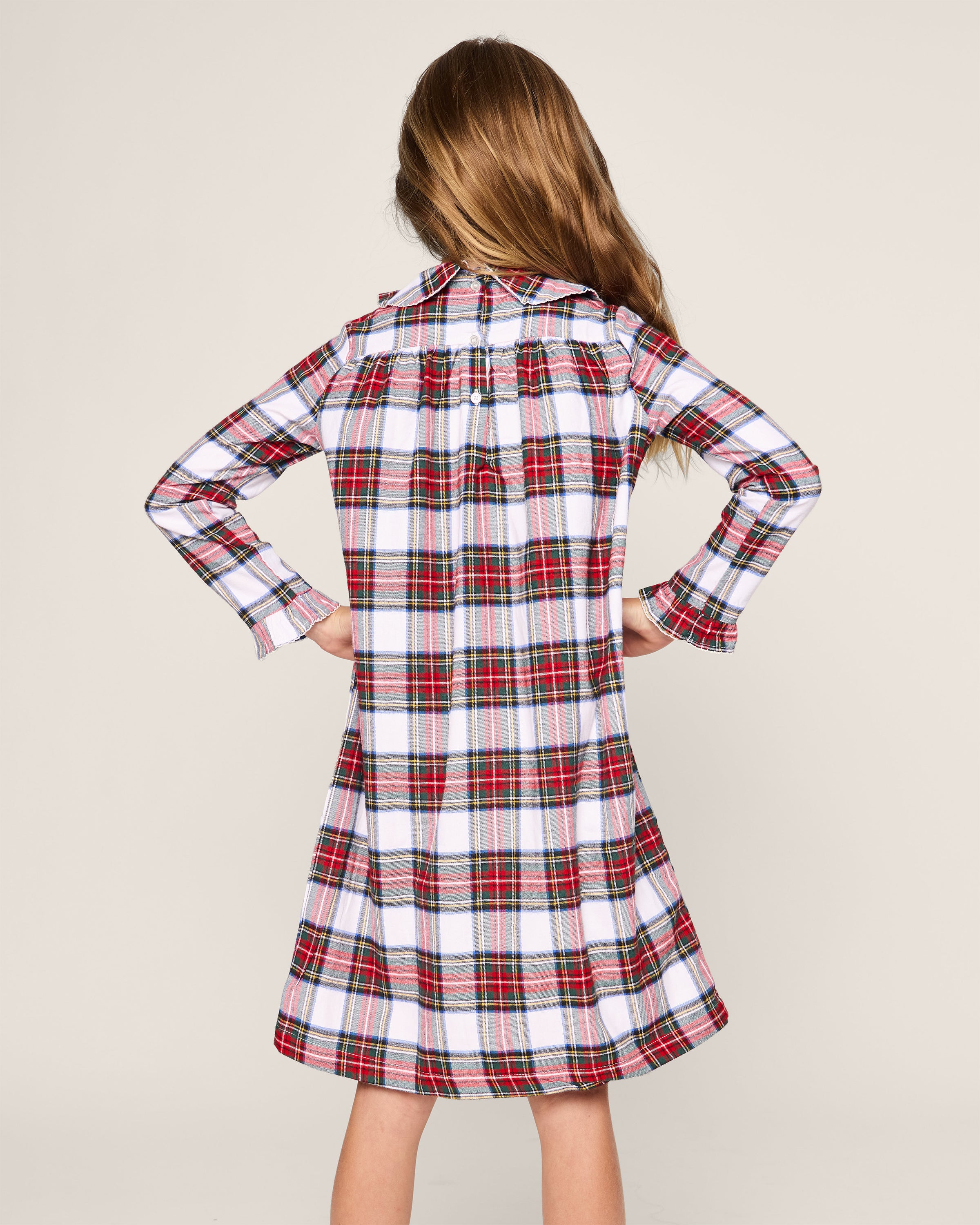 A young girl with long brown hair stands with hands on hips, wearing Petite Plumes Girls Brushed Cotton Eloise Nightgown in Balmoral Tartan. It features red, white, and black hues against a plain light beige background.
