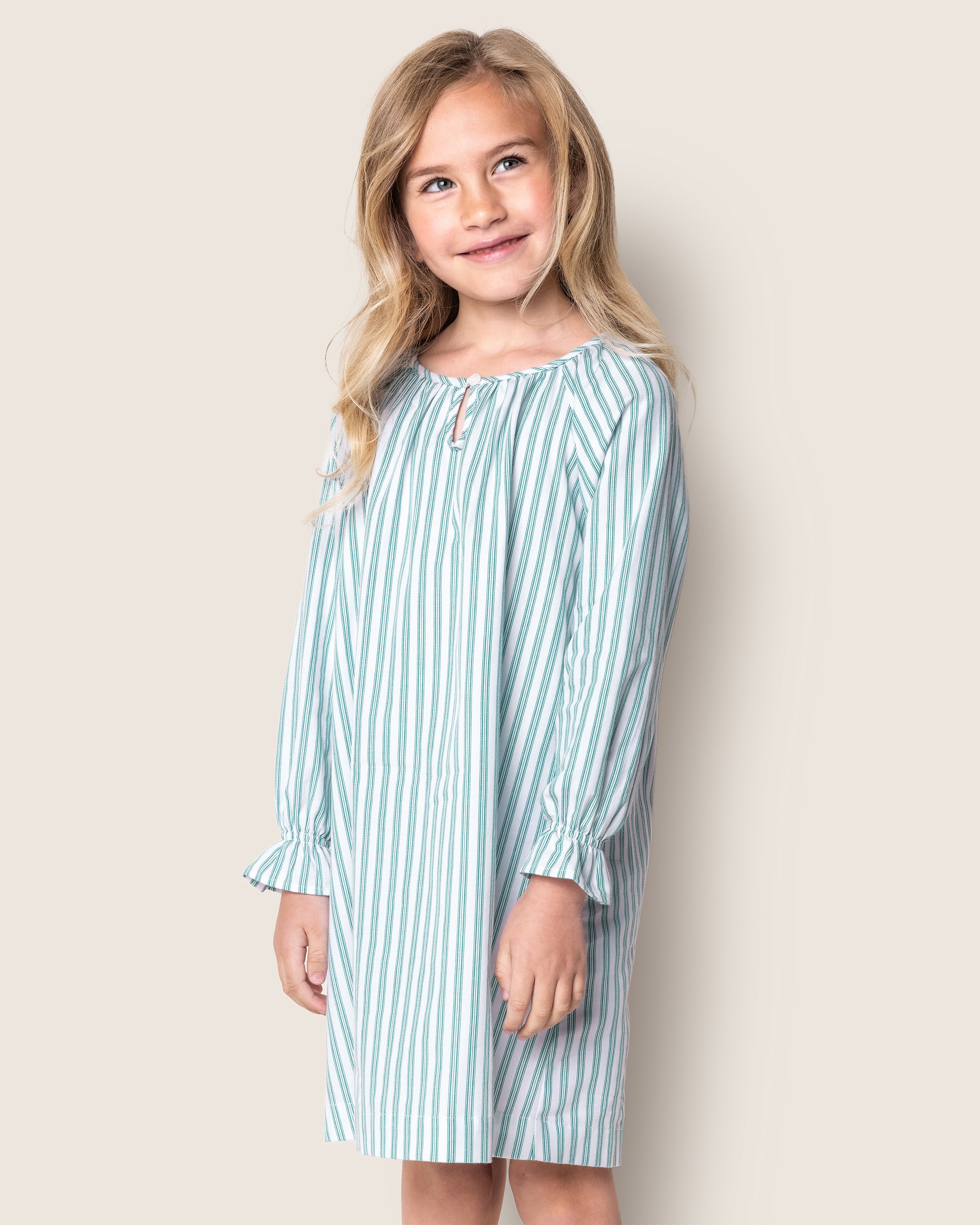 A young girl with long hair smiles while wearing the Girls Twill Delphine Nightgown in Emerald Ticking by Petite Plume. She stands against a plain background, looking slightly to the side.