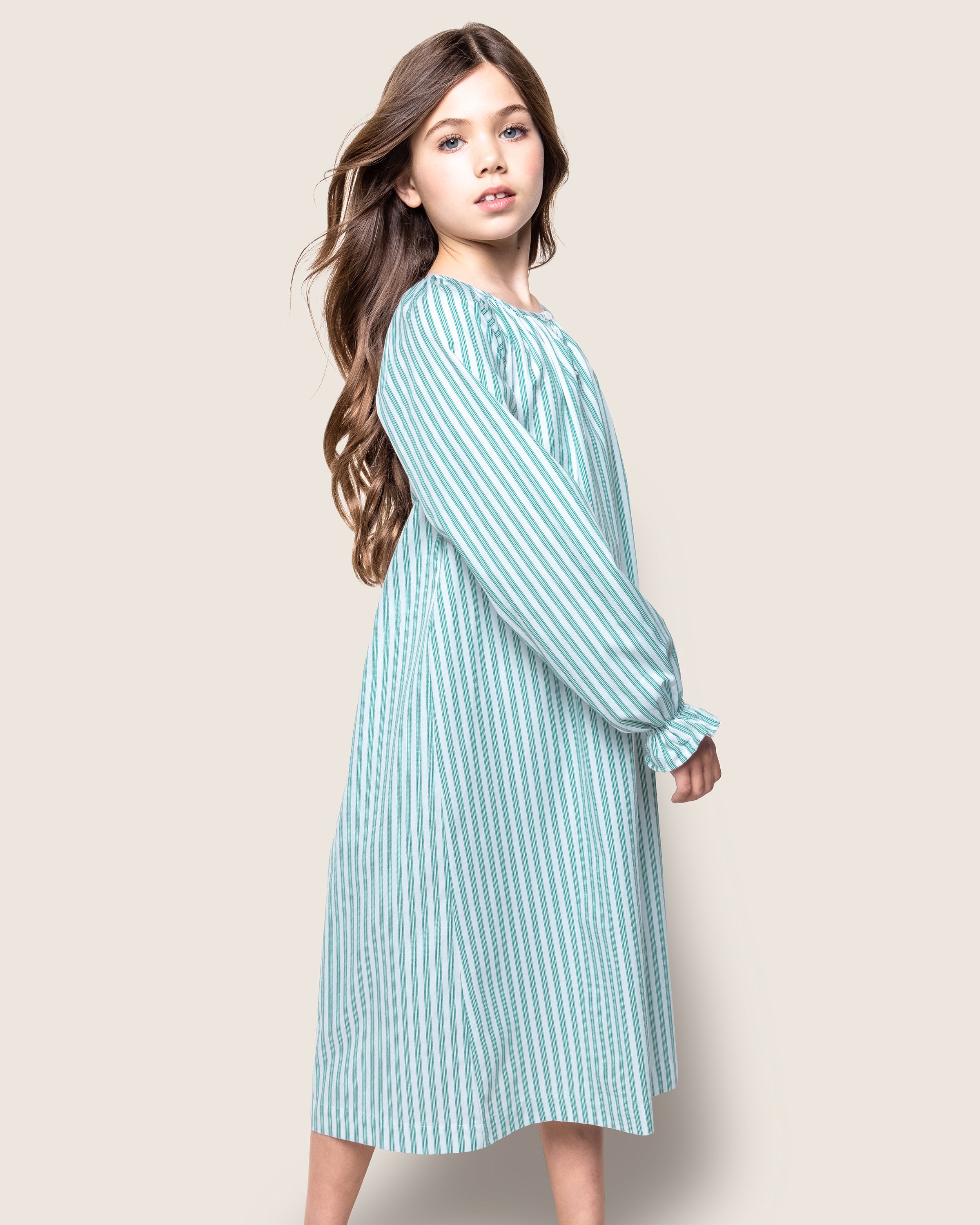 A young girl with long brown hair wears Petite Plumes Girls Twill Delphine Nightgown in Emerald Ticking, made of chemical-free flame-retardant fabric. She stands sideways, gazing at the camera with a neutral expression against a light gray background.