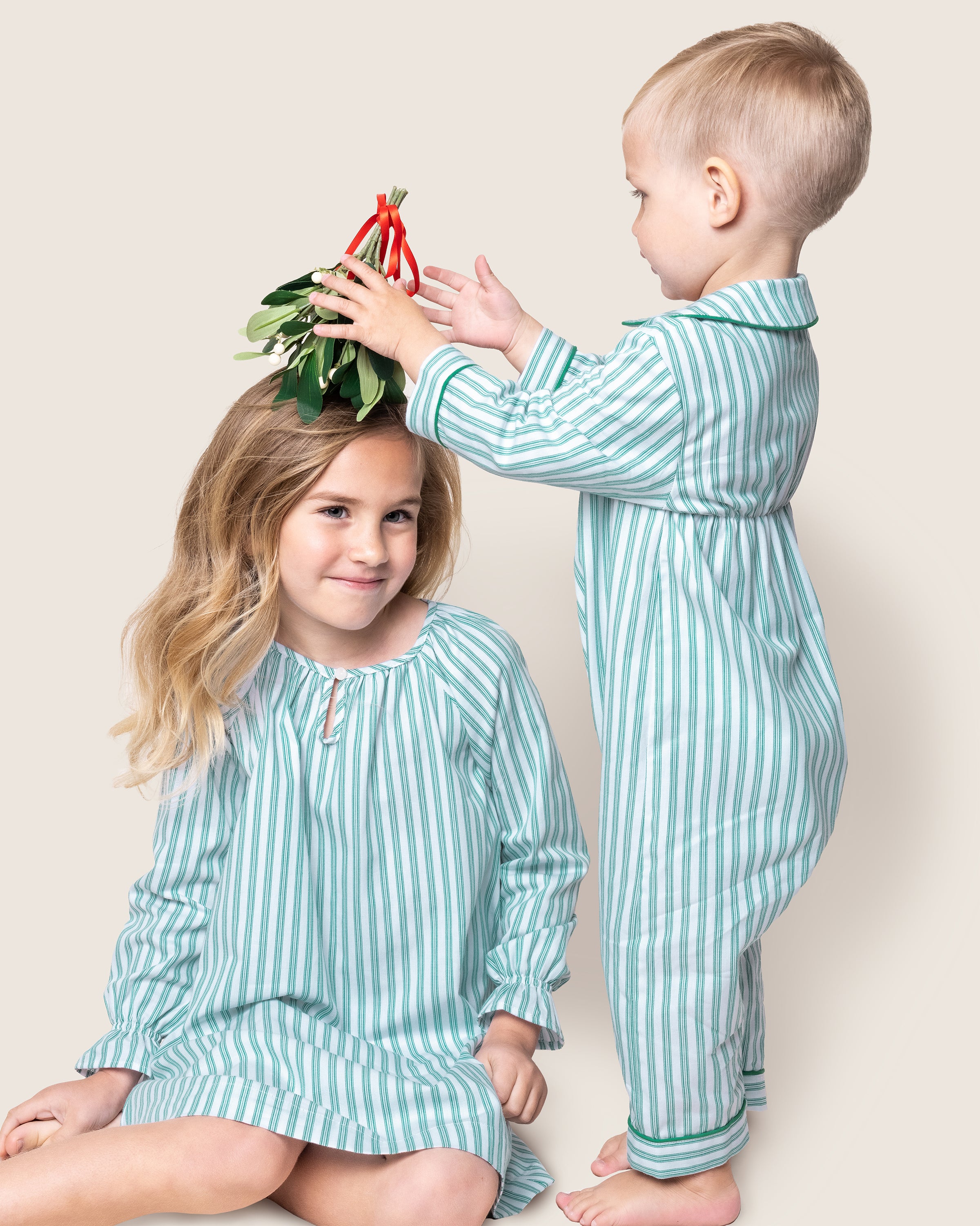 Two children in matching Petite Plume outfits are posed together. One smiles at the camera while the other playfully holds mistletoe overhead. Their charm shines against the light background, dressed in moisture-wicking attire similar to the Girls Twill Delphine Nightgown in Emerald Ticking.