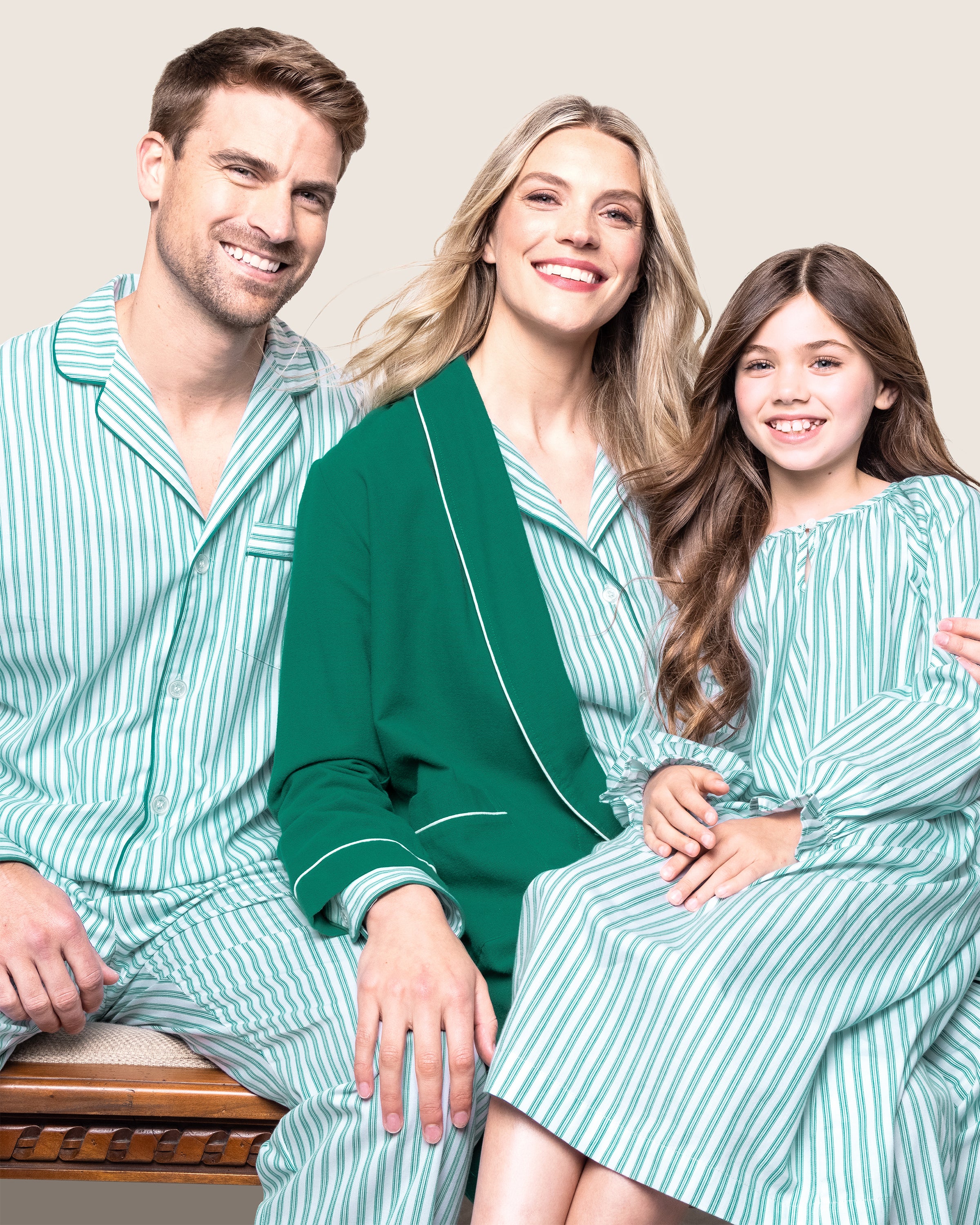 A smiling family of three is sitting happily on a bench. The man and child are wearing matching green and white striped pajamas in chemical-free flame-retardant fabric, while the woman wears a green robe. The brown-haired child is dressed in Petite Plumes Girls Twill Delphine Nightgown in Emerald Ticking.