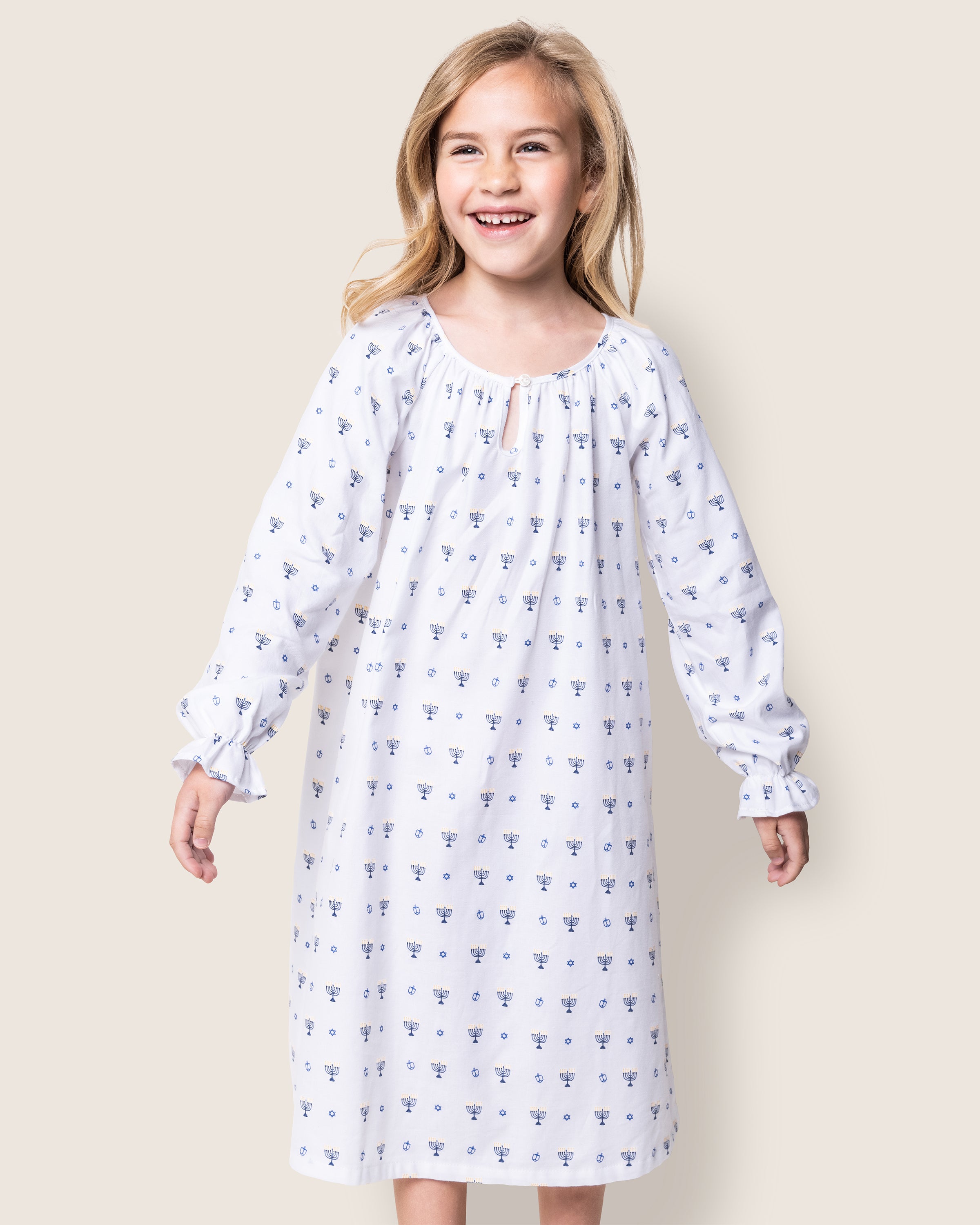 A young girl beams in her Girls Twill Delphine Nightgown in Happy Hanukkah by Petite Plume, with a small floral pattern capturing the essence of classic childrens sleepwear against a neutral backdrop highlighting her serene presence.