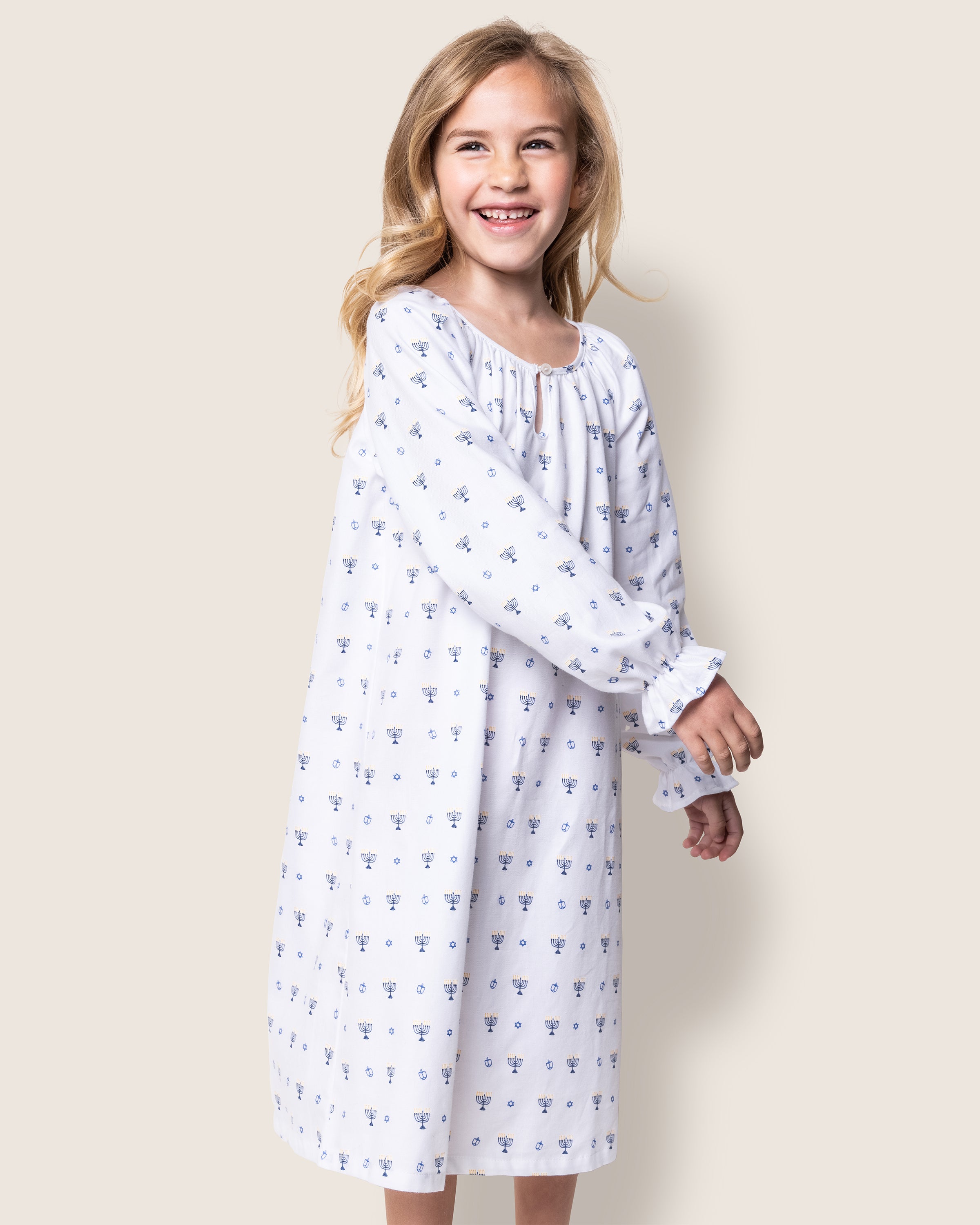 A young girl with long, blonde hair smiles while wearing Petite Plumes flame-retardant Girls Twill Delphine Nightgown in Happy Hanukkah—a white nightgown with small blue floral patterns. She stands against a plain, light background, looking slightly to her left.