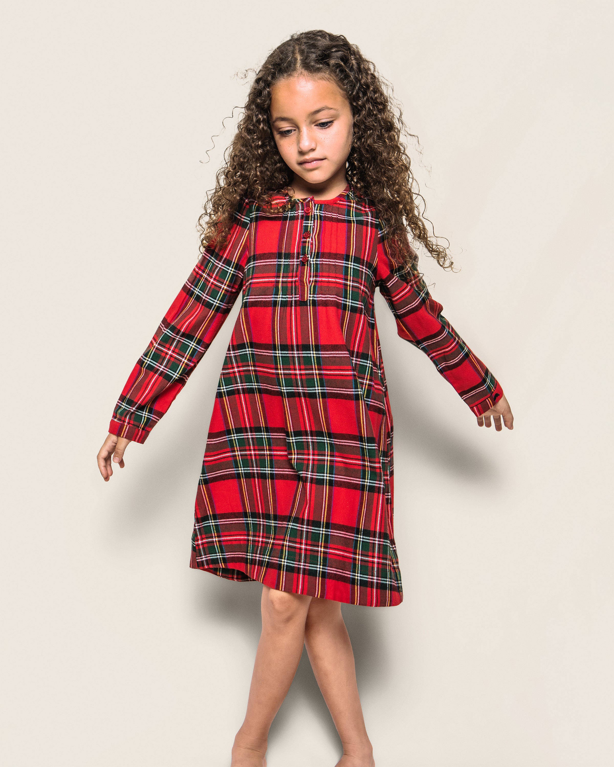 A young girl with curly hair is wearing Petite Plumes Girls Brushed Cotton Beatrice Nightgown in Imperial Tartan. She stands barefoot against a neutral background, looking down to the side with her arms slightly raised.
