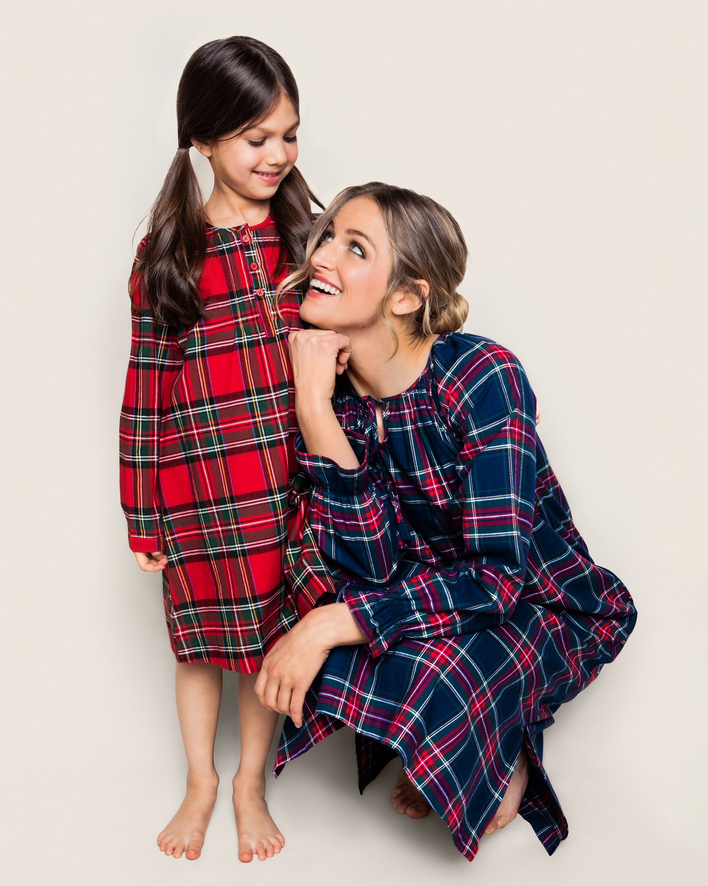 A woman crouches next to a young girl in pigtails, both smiling; the woman wears a blue long-sleeved Imperial Tartan plaid dress, and the girl wears Petite Plumes red Girls Brushed Cotton Beatrice Nightgown in Imperial Tartan as they joyfully gaze at each other against a plain background.