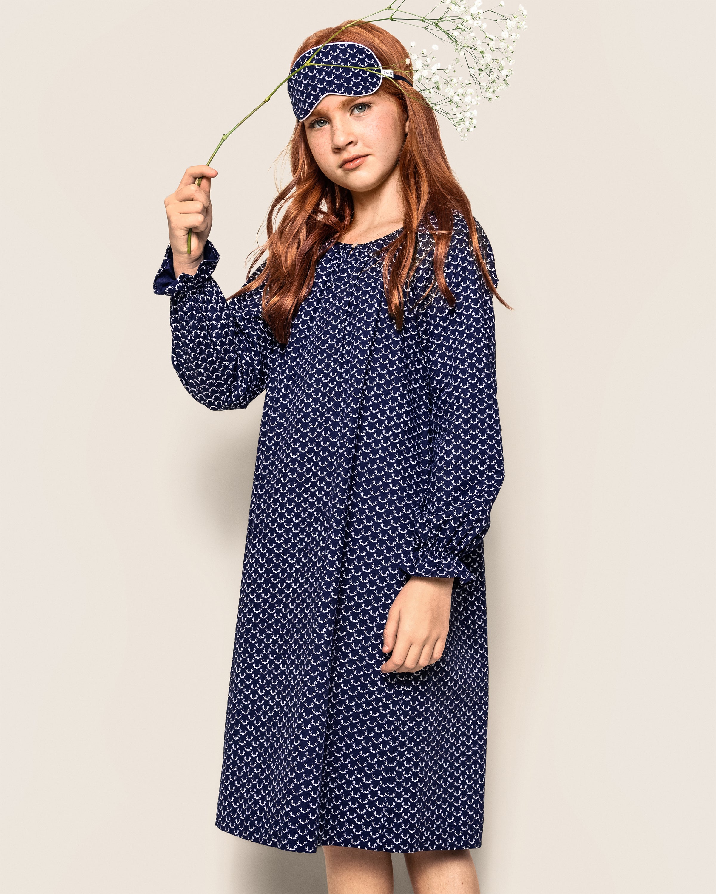 A young girl with long red hair stands in a Girls Twill Delphine Nightgown in Nordic Antlers by Petite Plume, with a matching sleep mask on her forehead. She holds a stem of white flowers, set against a plain beige background.