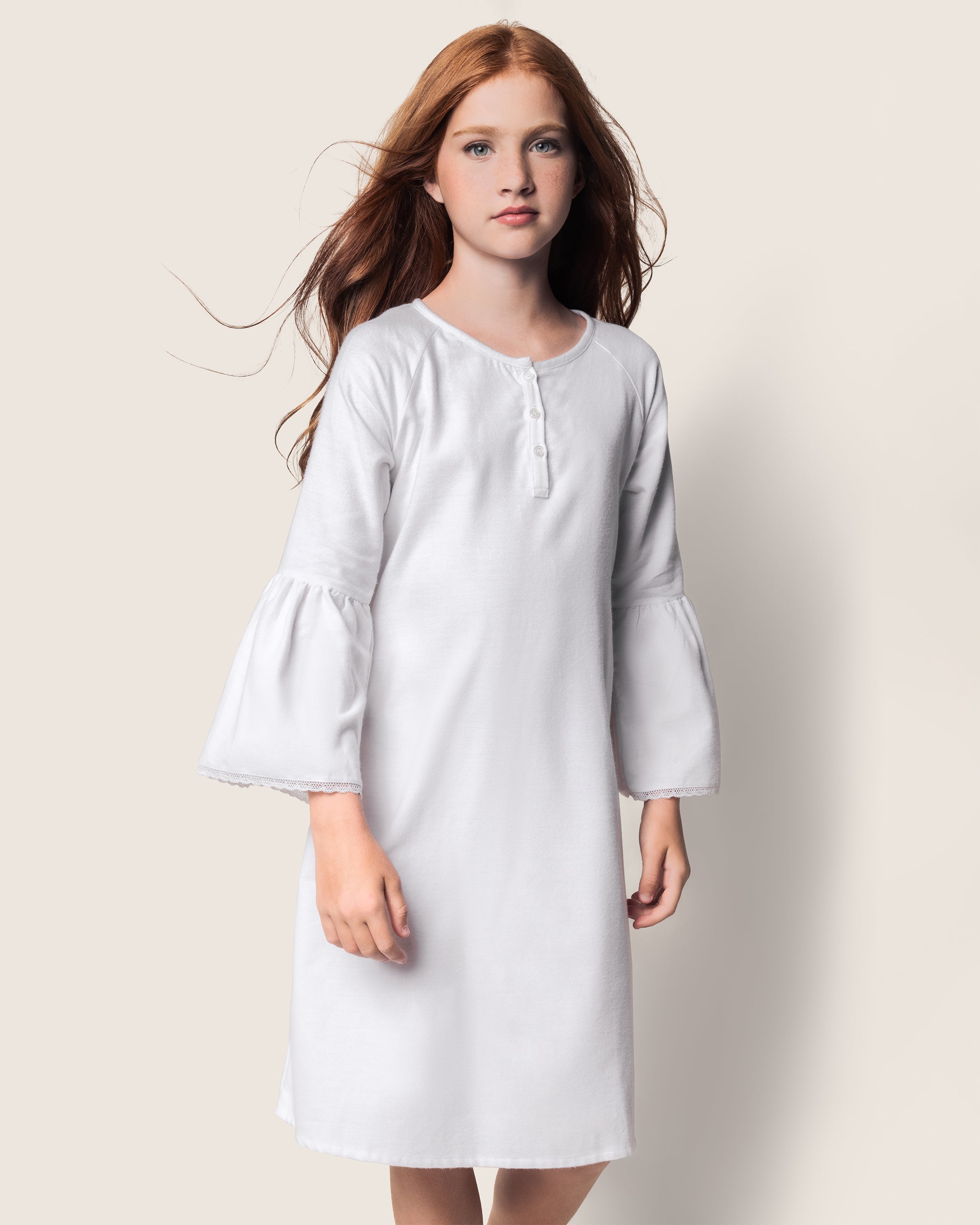 A girl with long red hair poses in Petite Plumes Girls Flannel Seraphine Nightgown in white, featuring bell sleeves and a buttoned neckline, set against a plain background.