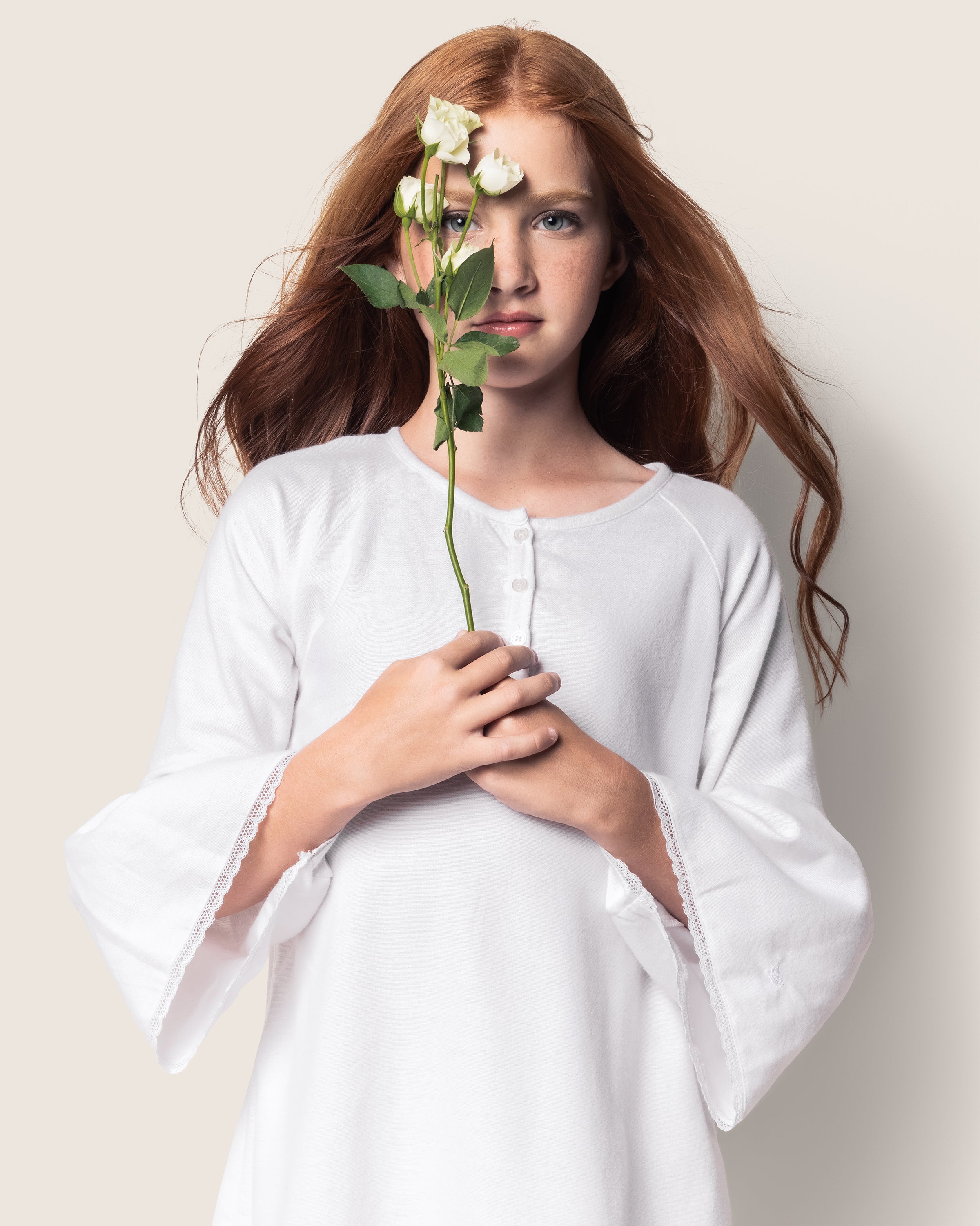 A person with long, wavy red hair holds a white flower in front of their face while wearing Petite Plumes Girls Flannel Seraphine Nightgown in White. They stand against a neutral background.